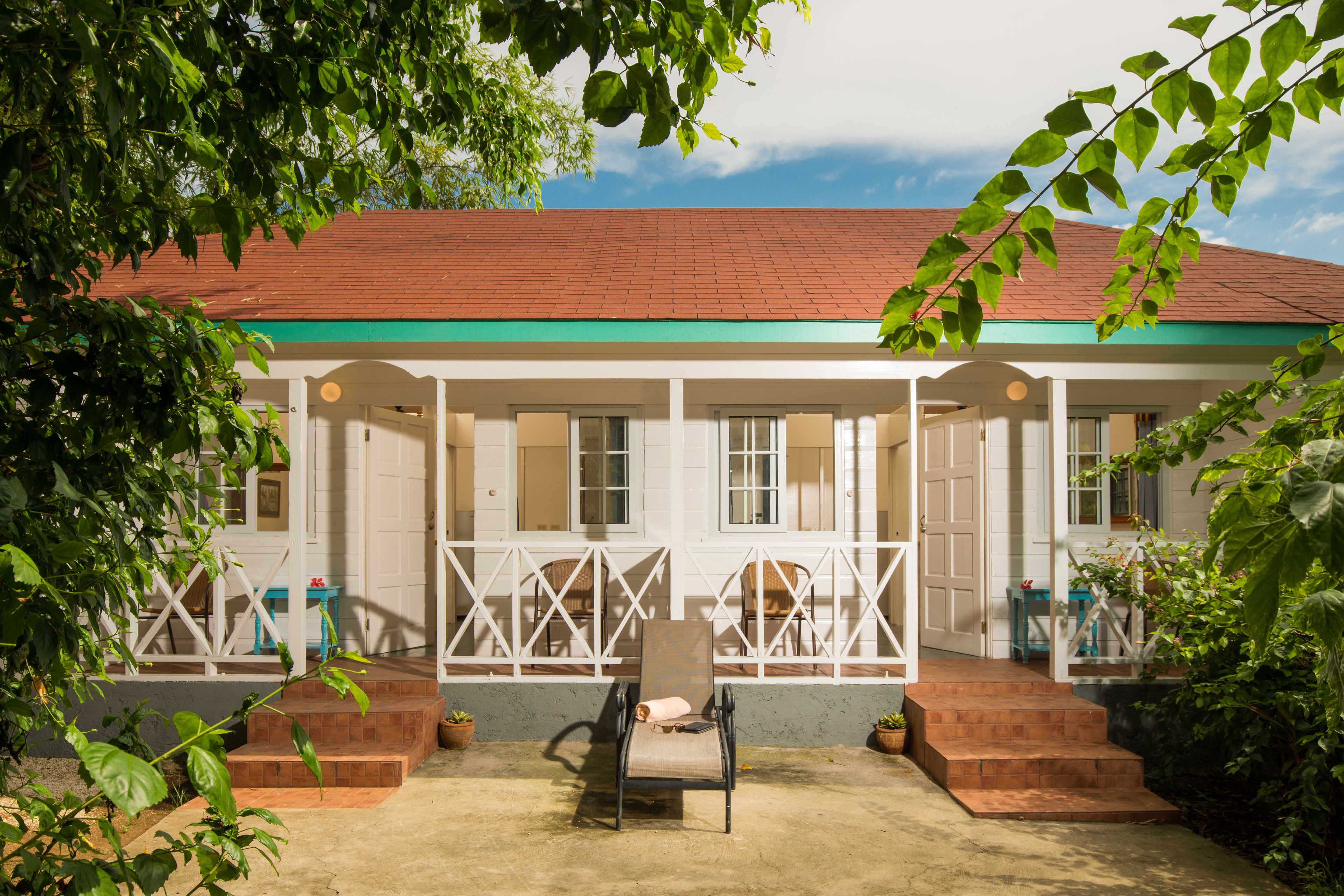 A two-story building with a red roof and white walls, featuring a covered porch with chairs and potted plants.