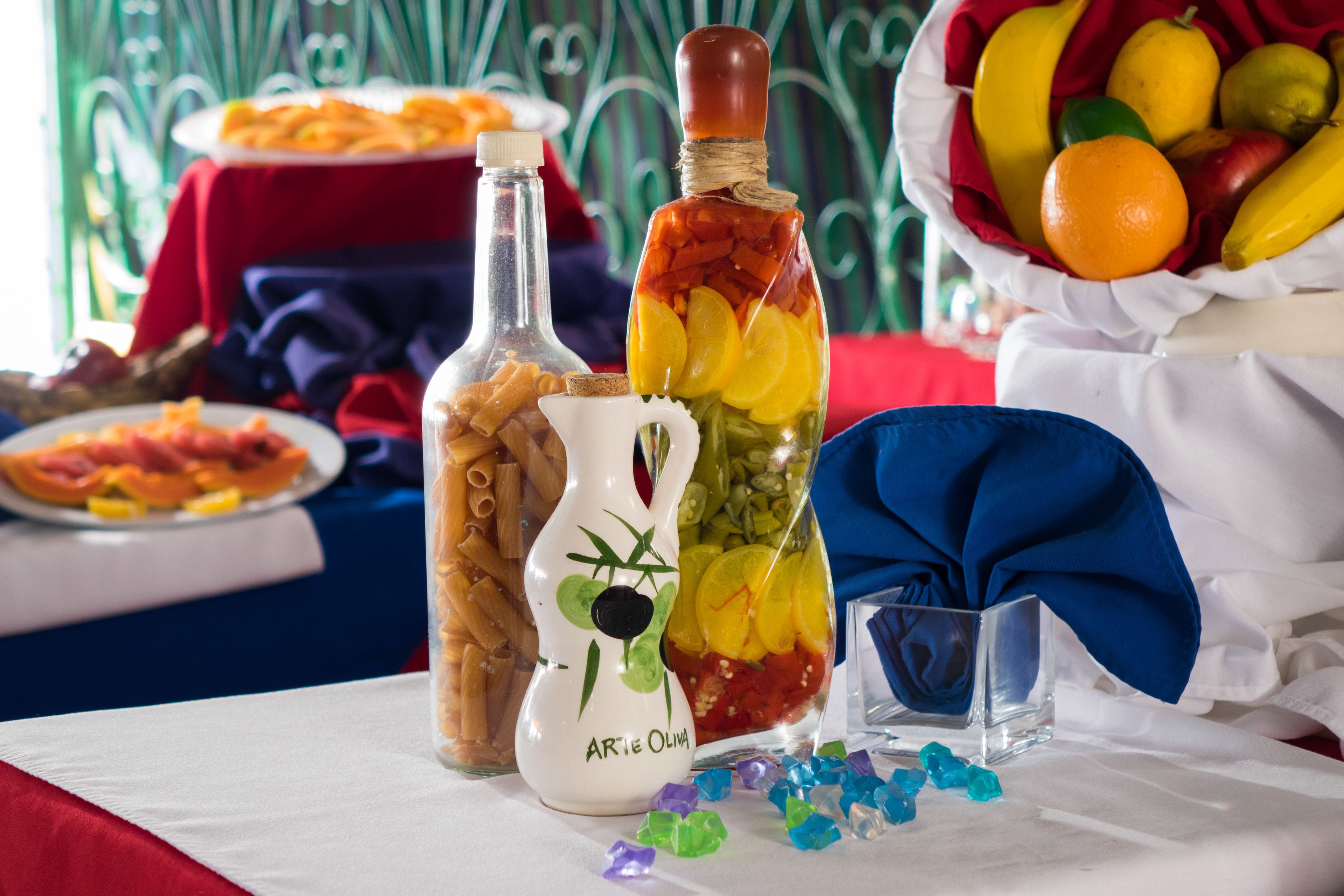 A table with various food items, including a bottle of olives, a pitcher, and a basket of fruits.