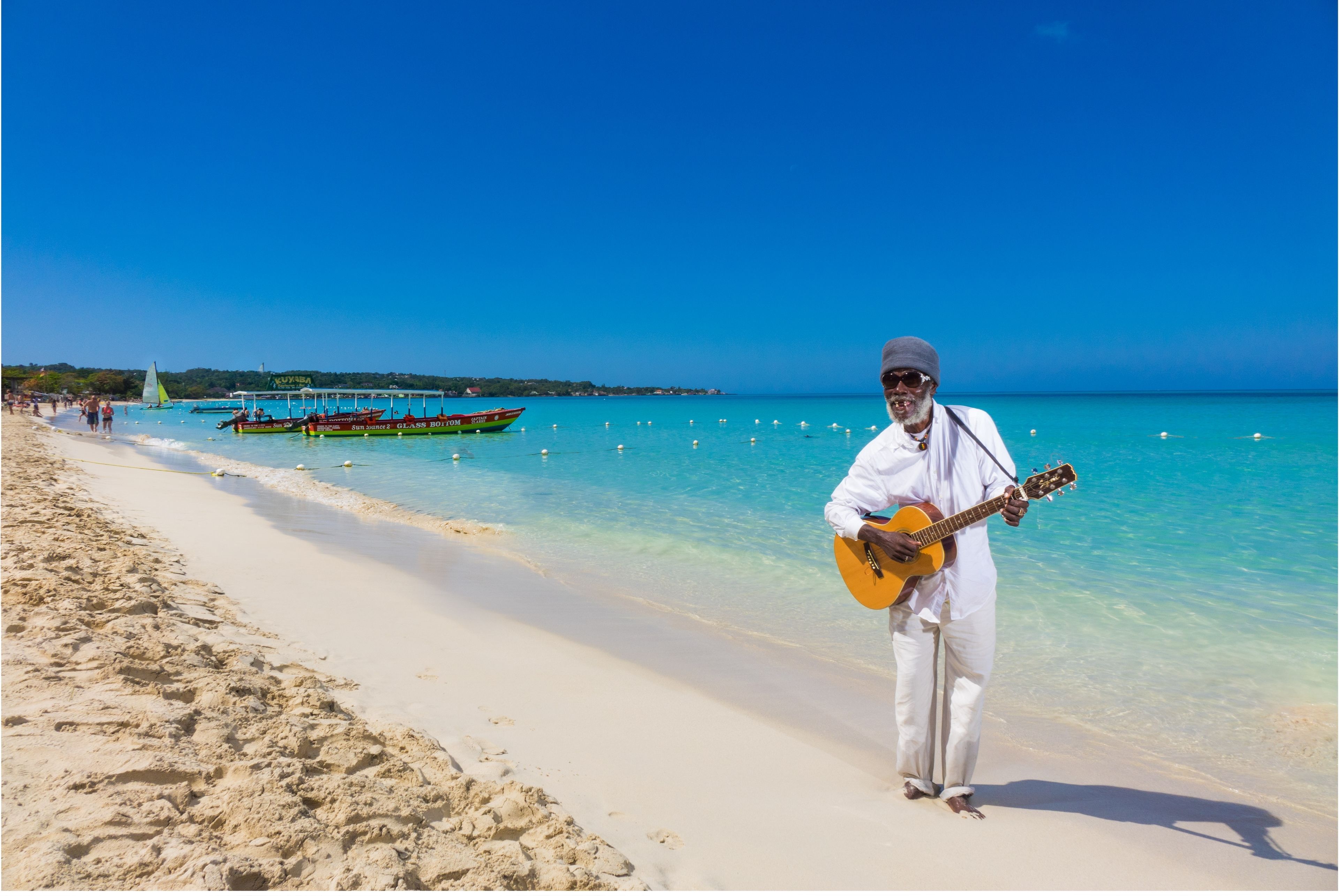 Una spiaggia con acque turchesi cristalline, un uomo che suona la chitarra e barche sullo sfondo.