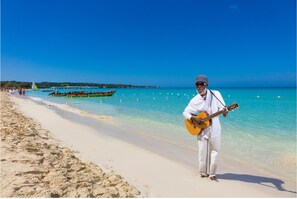A beach with clear turquoise water, a man playing a guitar, and boats in the background.