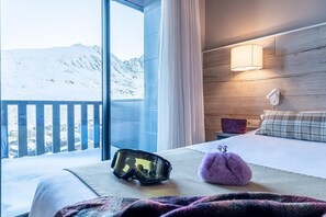 A hotel room with a large window, a bed, a nightstand, and a ski helmet on the bed. - Hotel Caribou (Pas de la Casa)