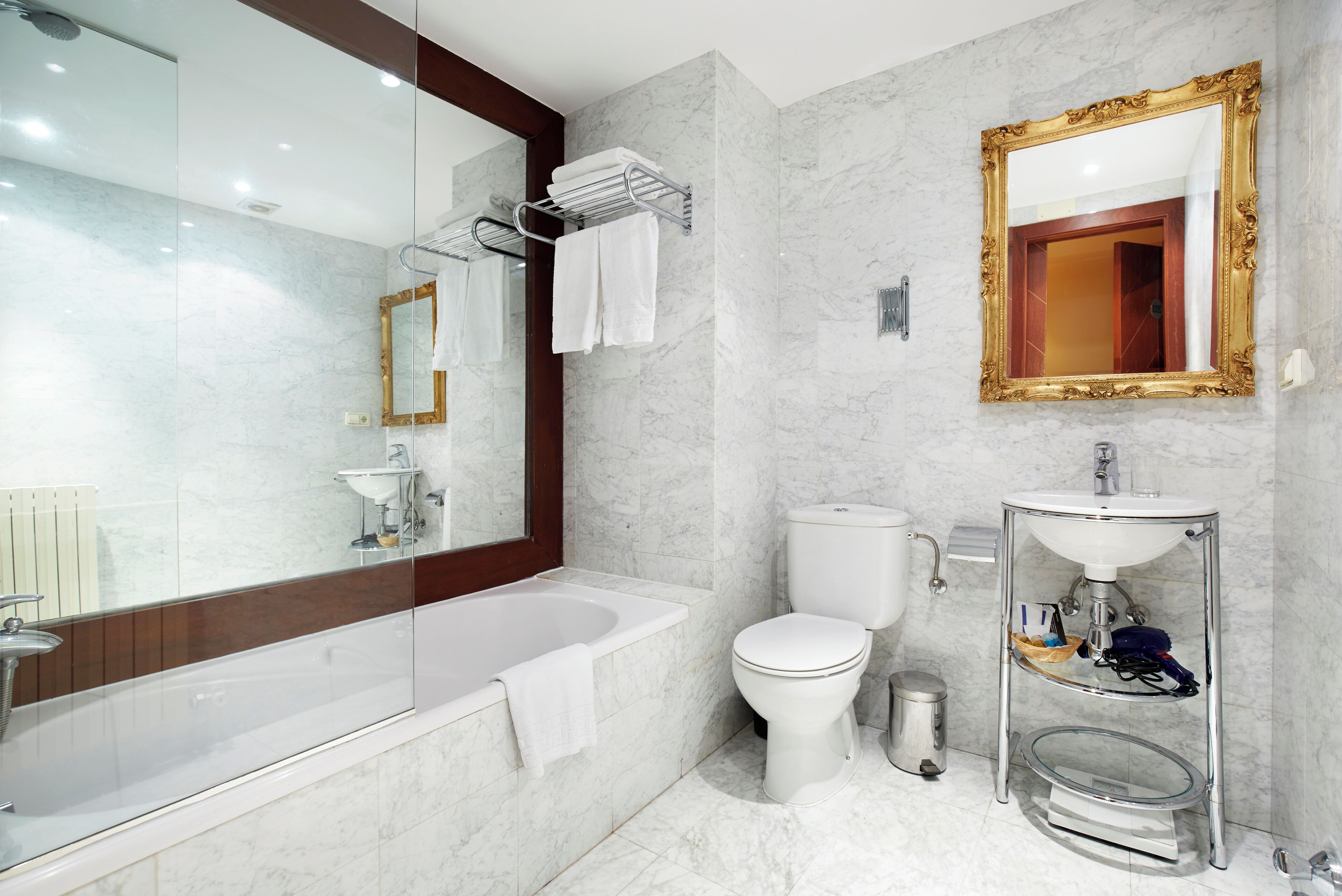 A modern bathroom with a glass-enclosed bathtub, a white toilet, a sink with a glass shelf, and a large mirror with a gold frame.