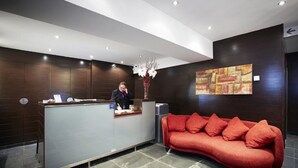 Uma área de recepção com balcão, recepcionista, um sofá vermelho e uma parede com pintura geométrica.