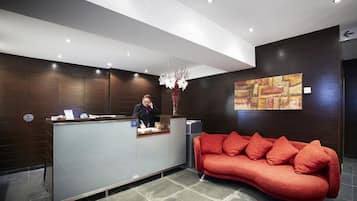 Uma área de recepção com balcão, recepcionista, sofá vermelho e uma parede com pintura abstrata.