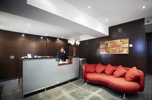 Uma área de recepção com balcão, recepcionista, um sofá vermelho e uma parede com pintura geométrica.