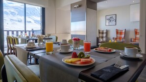 Cenário de café da manhã com vista para montanhas cobertas de neve, mesa com pratos de frutas, café e suco, e um ambiente de estar aconchegante.