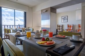 A breakfast setting with a view of snow-covered mountains, a table with plates of fruit, coffee, and juice, and a cozy seating arrangement. - Hotel Caribou (Pas de la Casa)