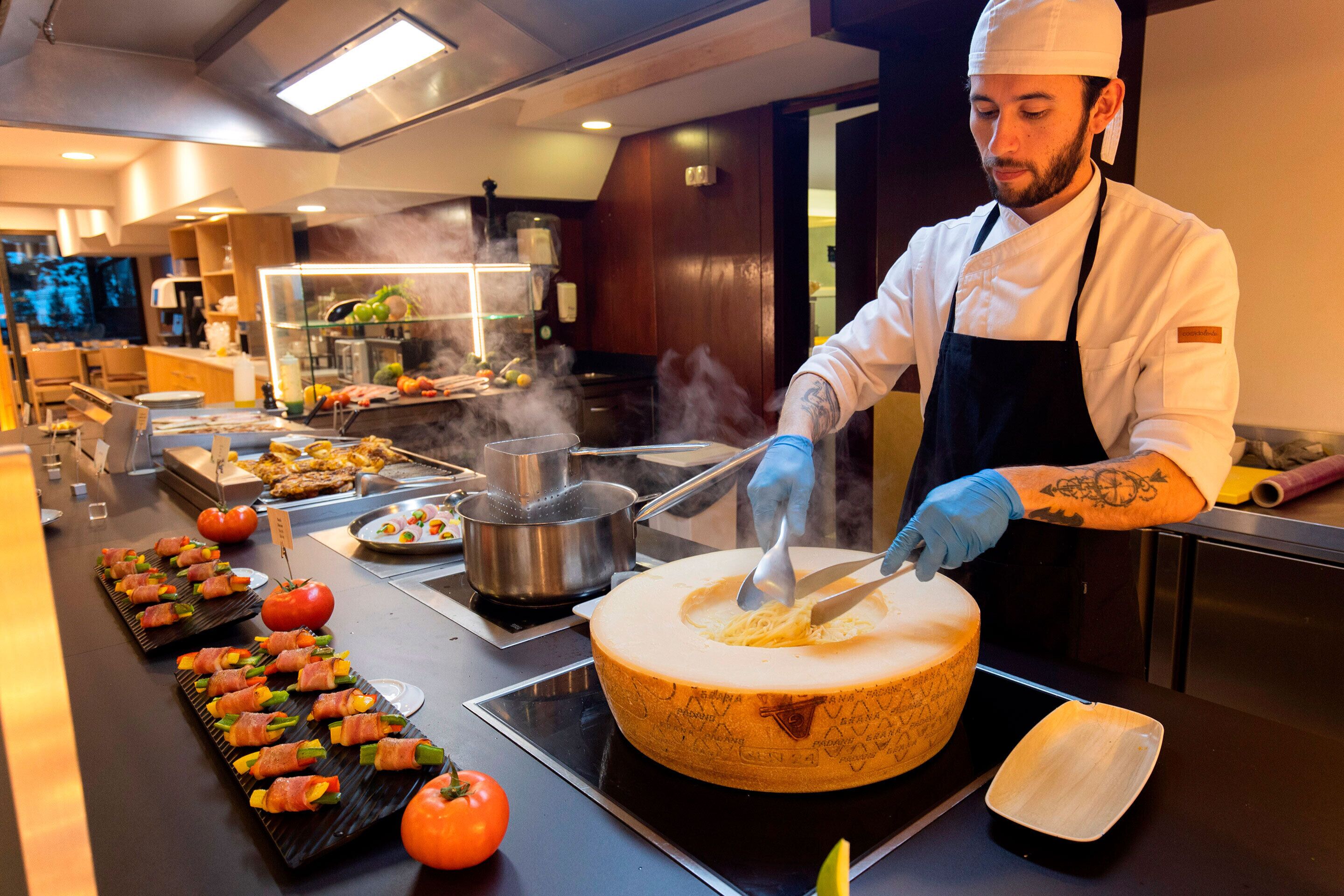 A chef preparing food in a kitchen with a buffet setup, including a large cheese wheel and various dishes.