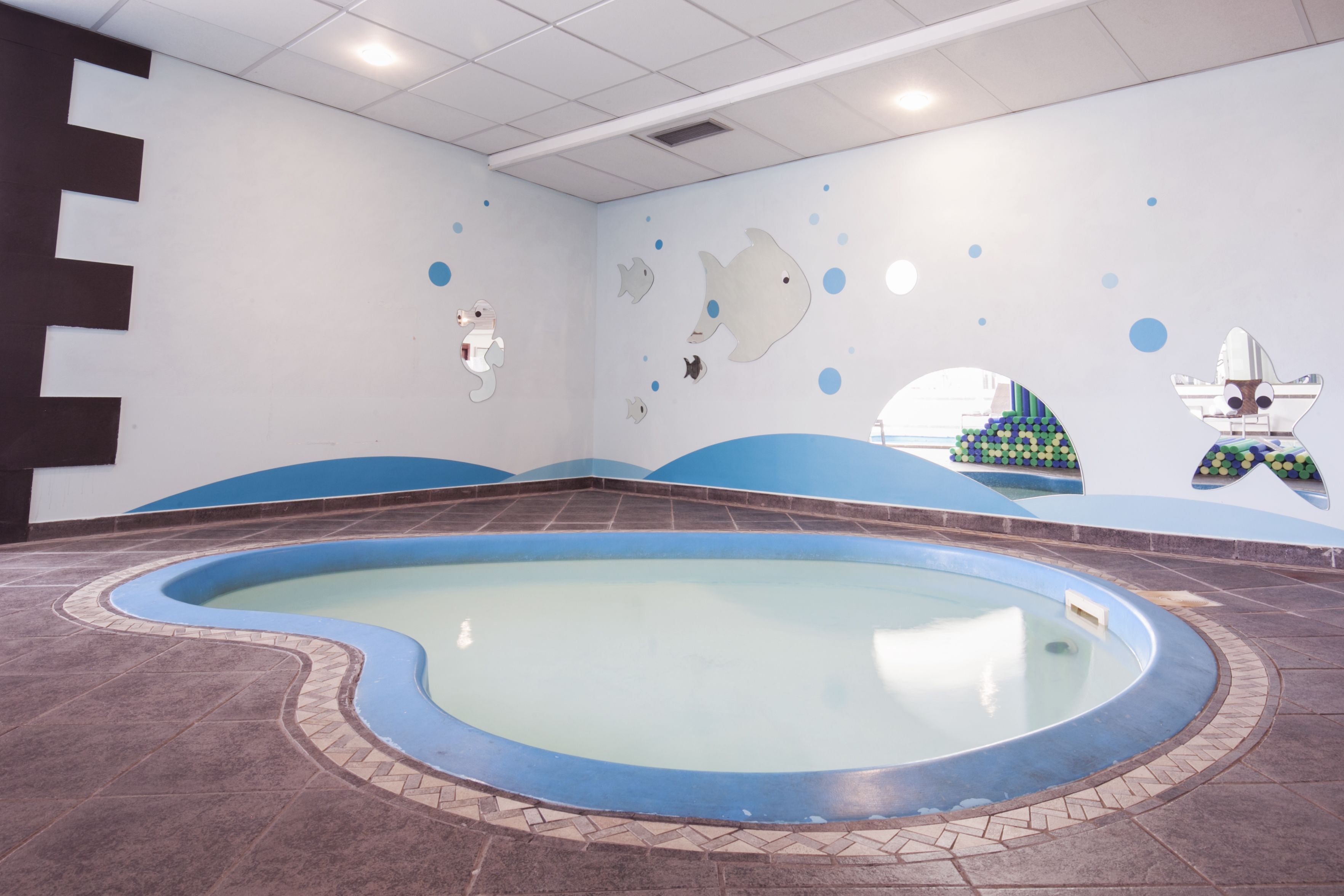 Una piscina con diseño azul y blanco, rodeada de piso y paredes con patrón de olas y decoraciones de peces.