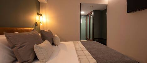 A hotel room with a large bed, a flat-screen TV mounted on the wall, and a hallway leading to another room.