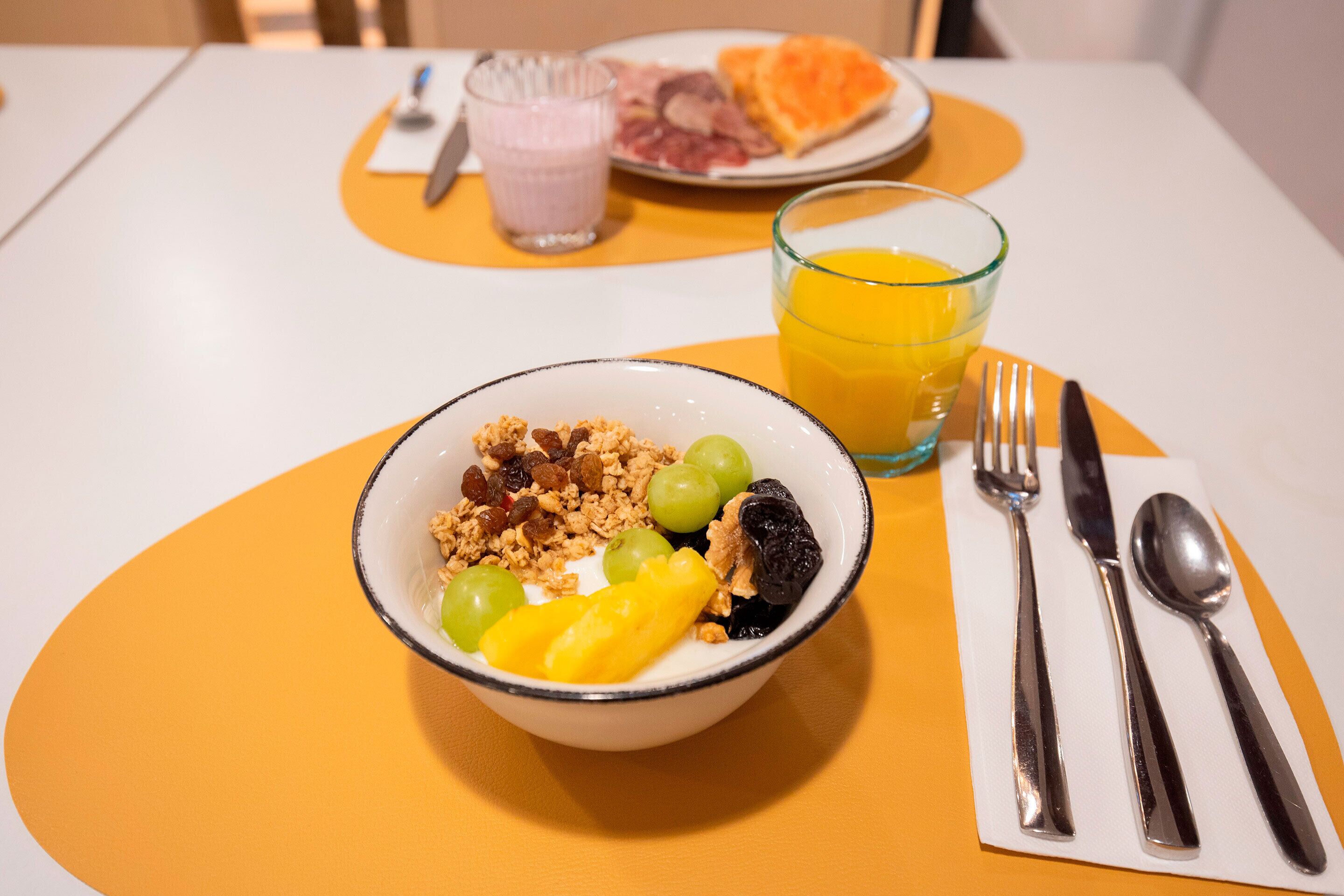 Una mesa con un tazón de cereales, frutas y un vaso de jugo de naranja.