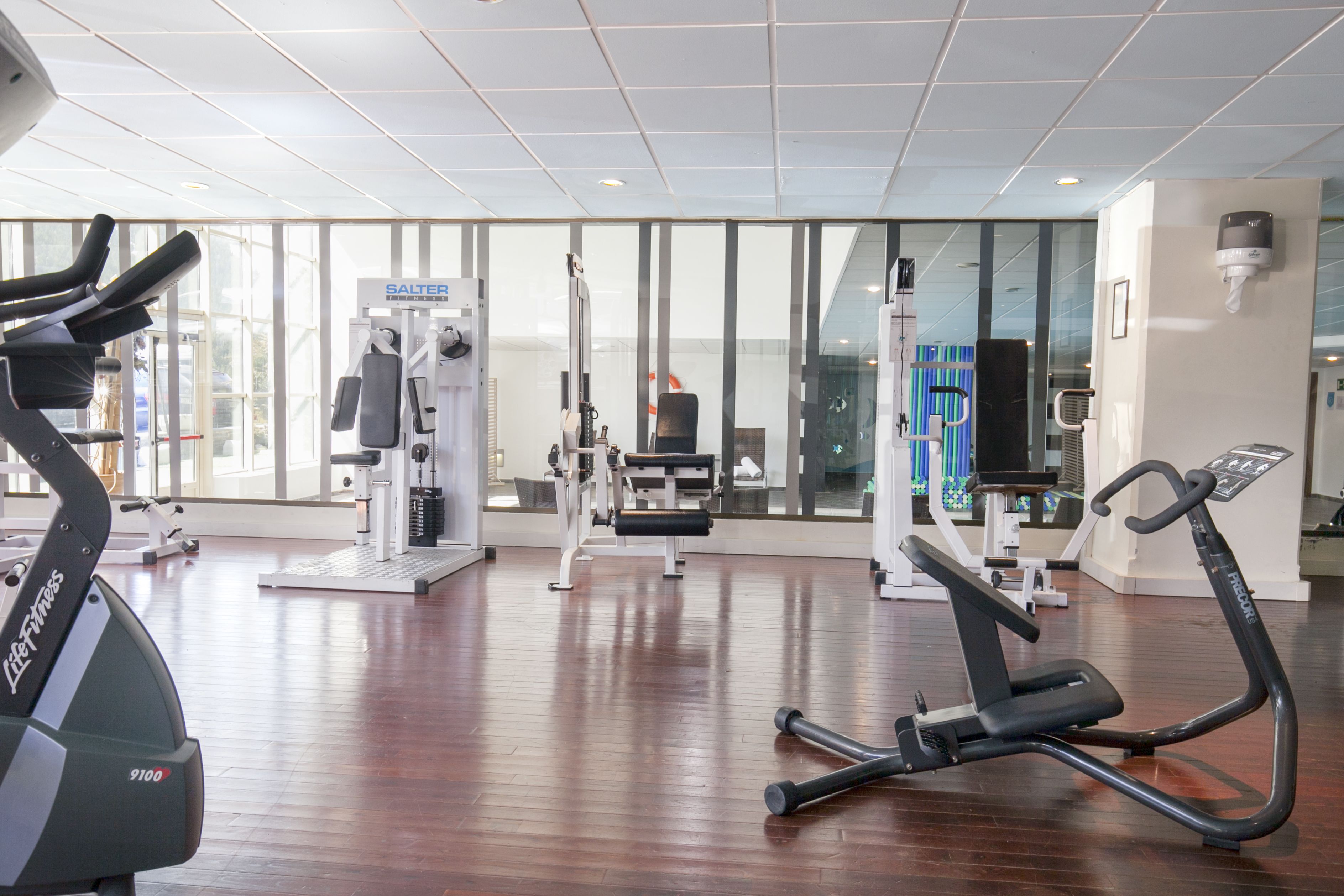 A gym with cardio machines, weight training equipment, and a large window providing natural light.