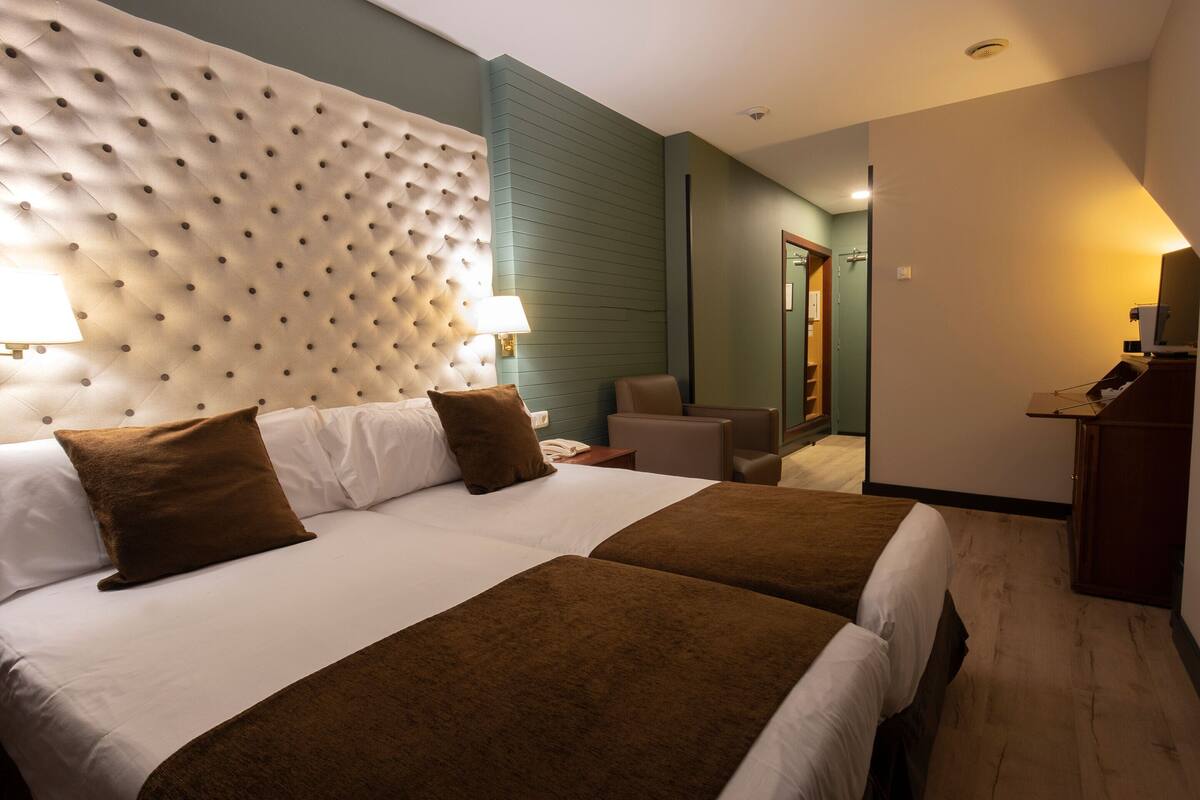 A hotel room with two beds, a tufted headboard, a desk with a lamp, and a small seating area.