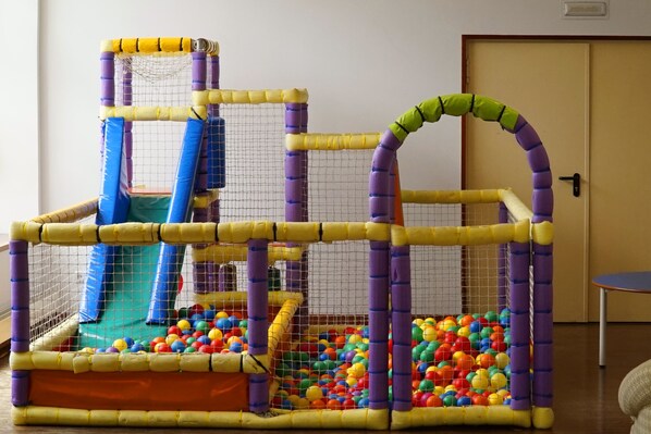Zona de juegos con piscina de pelotas y un tobogán, delimitada por una red con postes morados y amarillos.