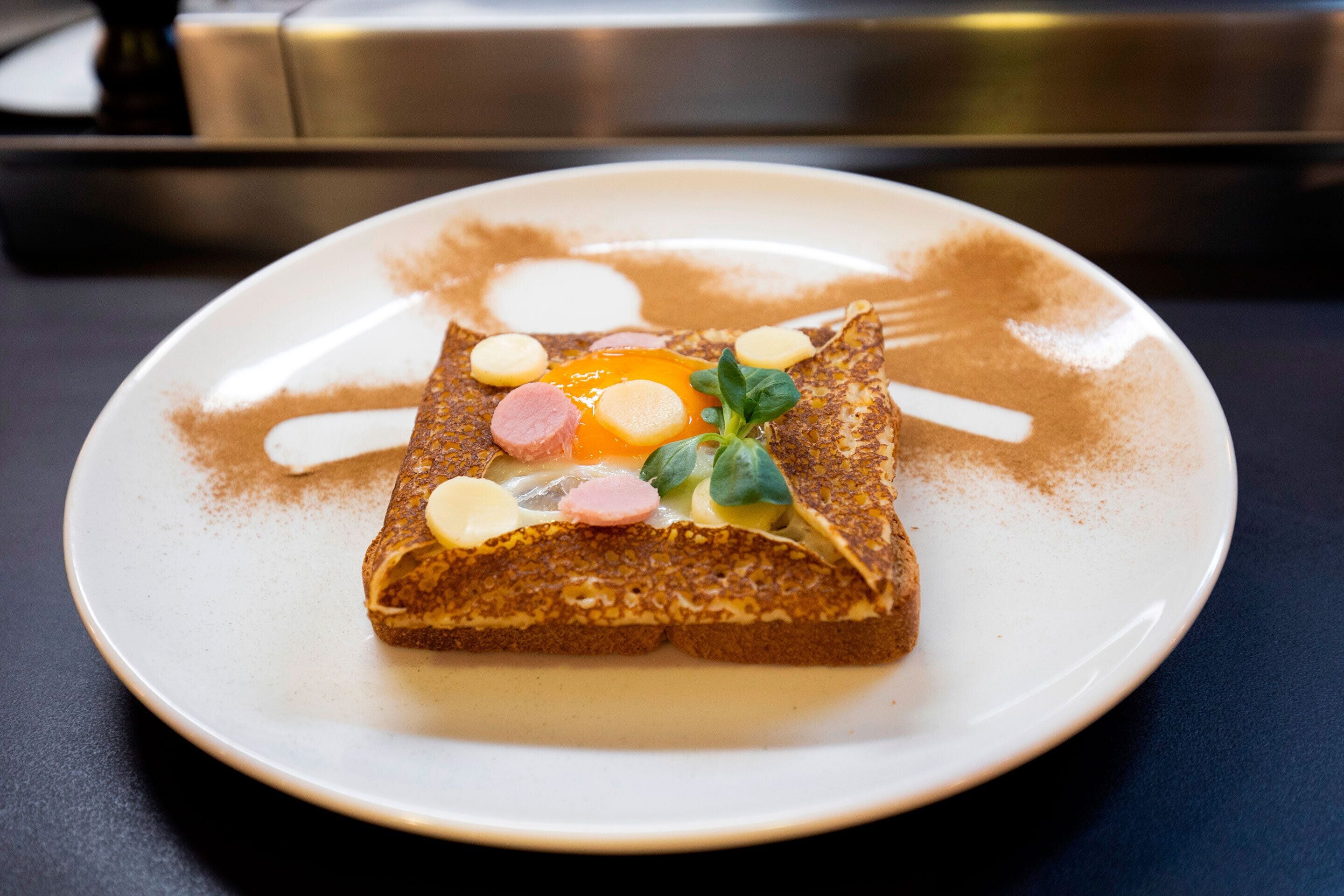 A plate with a crepe topped with ham, cheese, and a fried egg, garnished with herbs, on a white plate with a coffee stain.