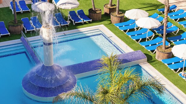 Una piscina con una fuente, rodeada de sillas de descanso azules y sombrillas blancas.