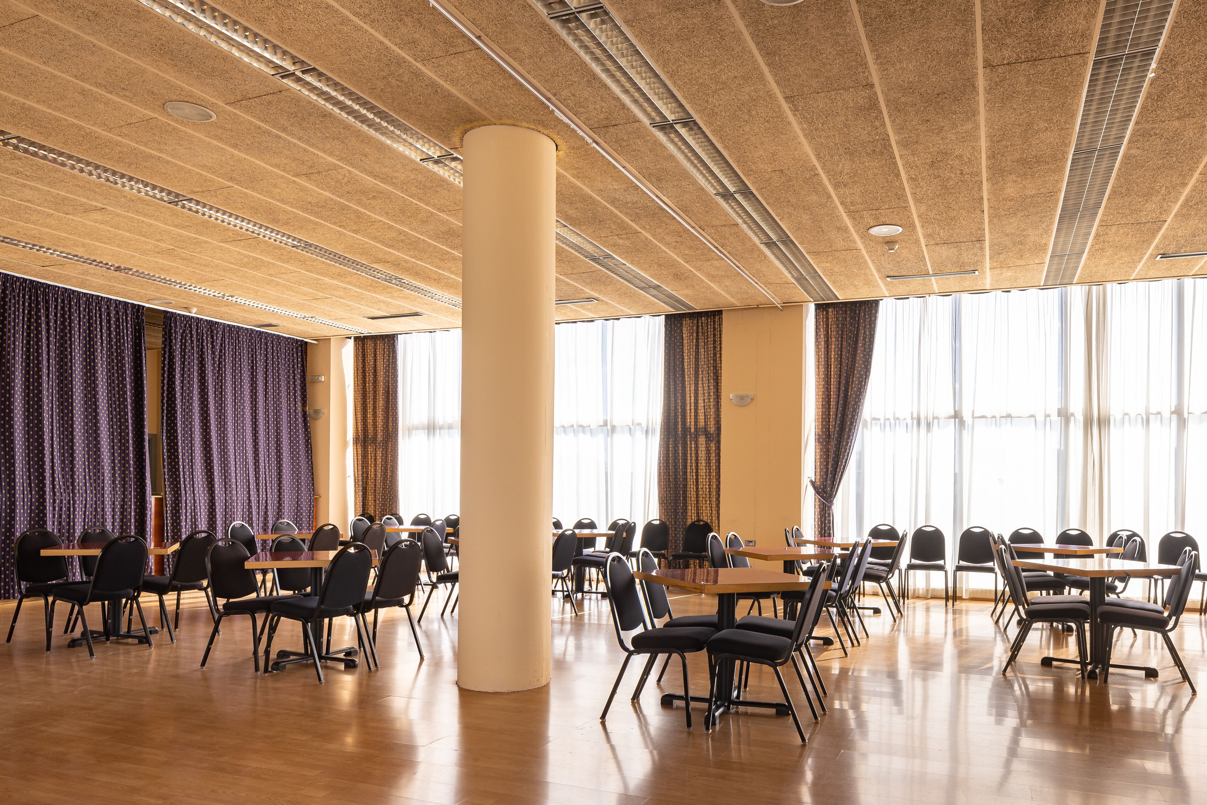 A spacious conference room with multiple tables and chairs, large windows with curtains, and a high ceiling with visible ductwork.