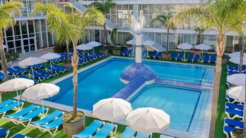A pool area with blue lounge chairs and white umbrellas, surrounded by palm trees and a modern building with balconies.