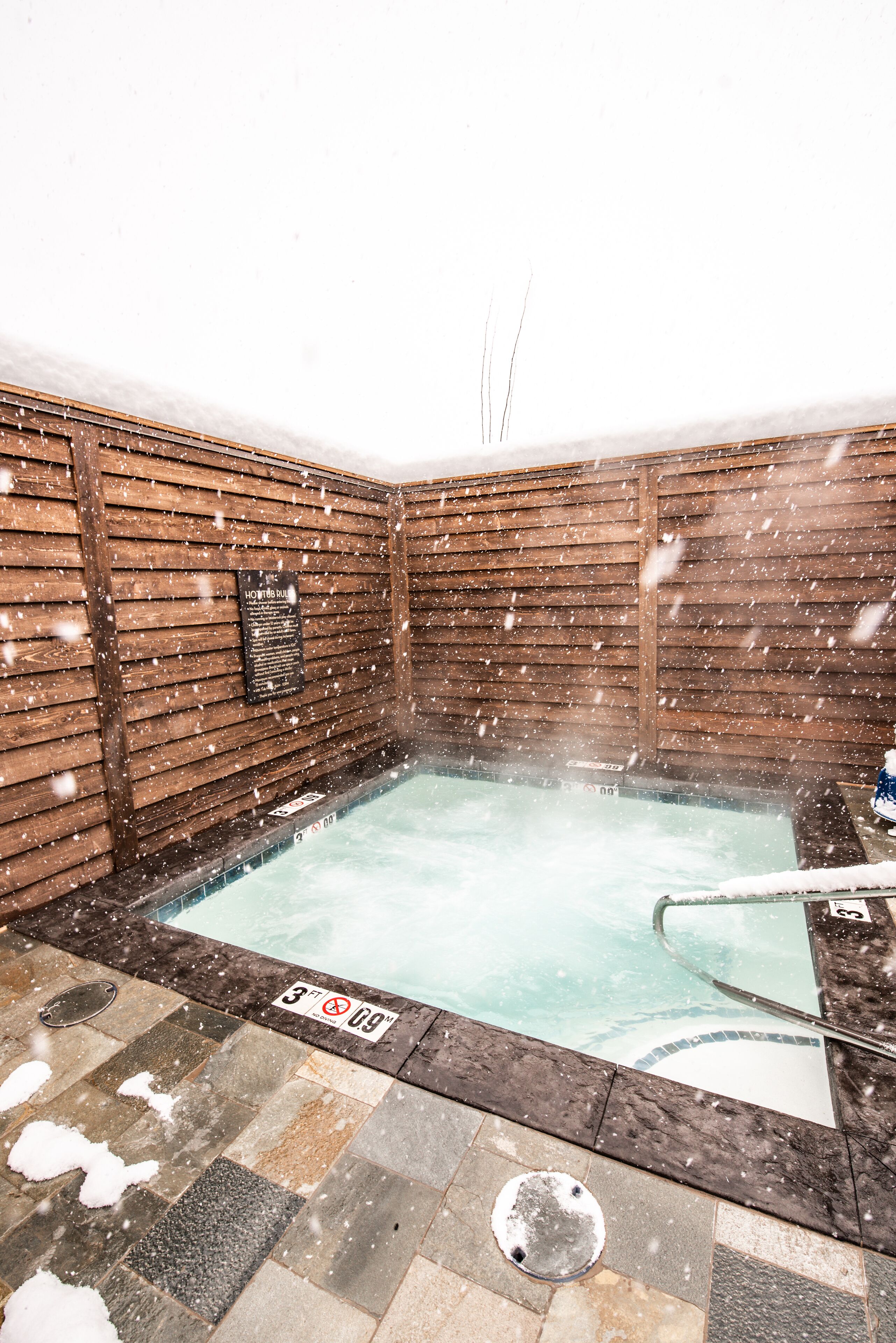 A hot tub with a wooden wall and a sign that reads "HOT TUB". Snow is falling outside.