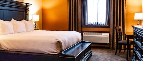 A hotel room with a large bed, a desk with a chair, an air conditioning unit, and a window with curtains.