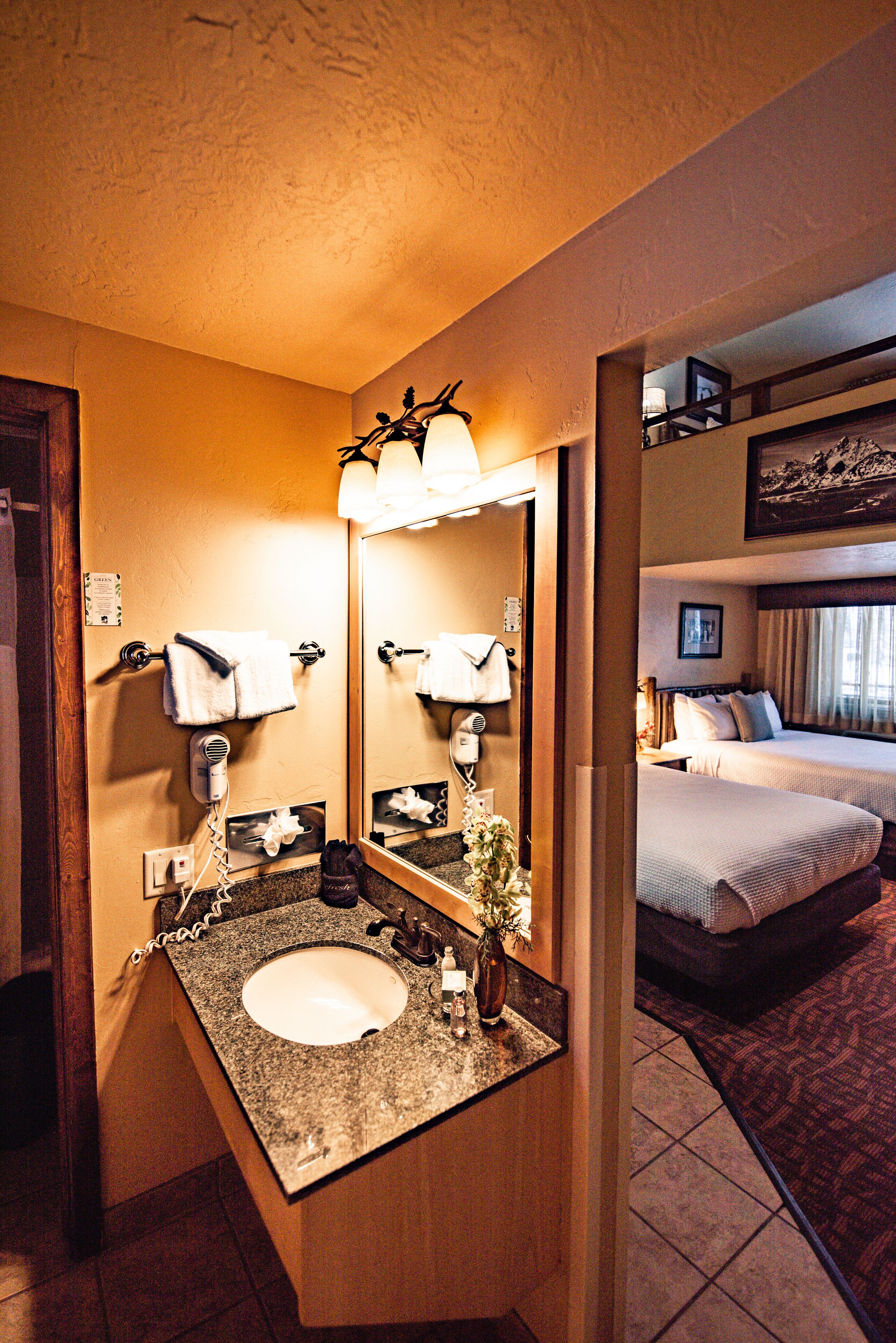 A hotel bathroom with a sink, mirror, and a bed visible through an adjacent room.