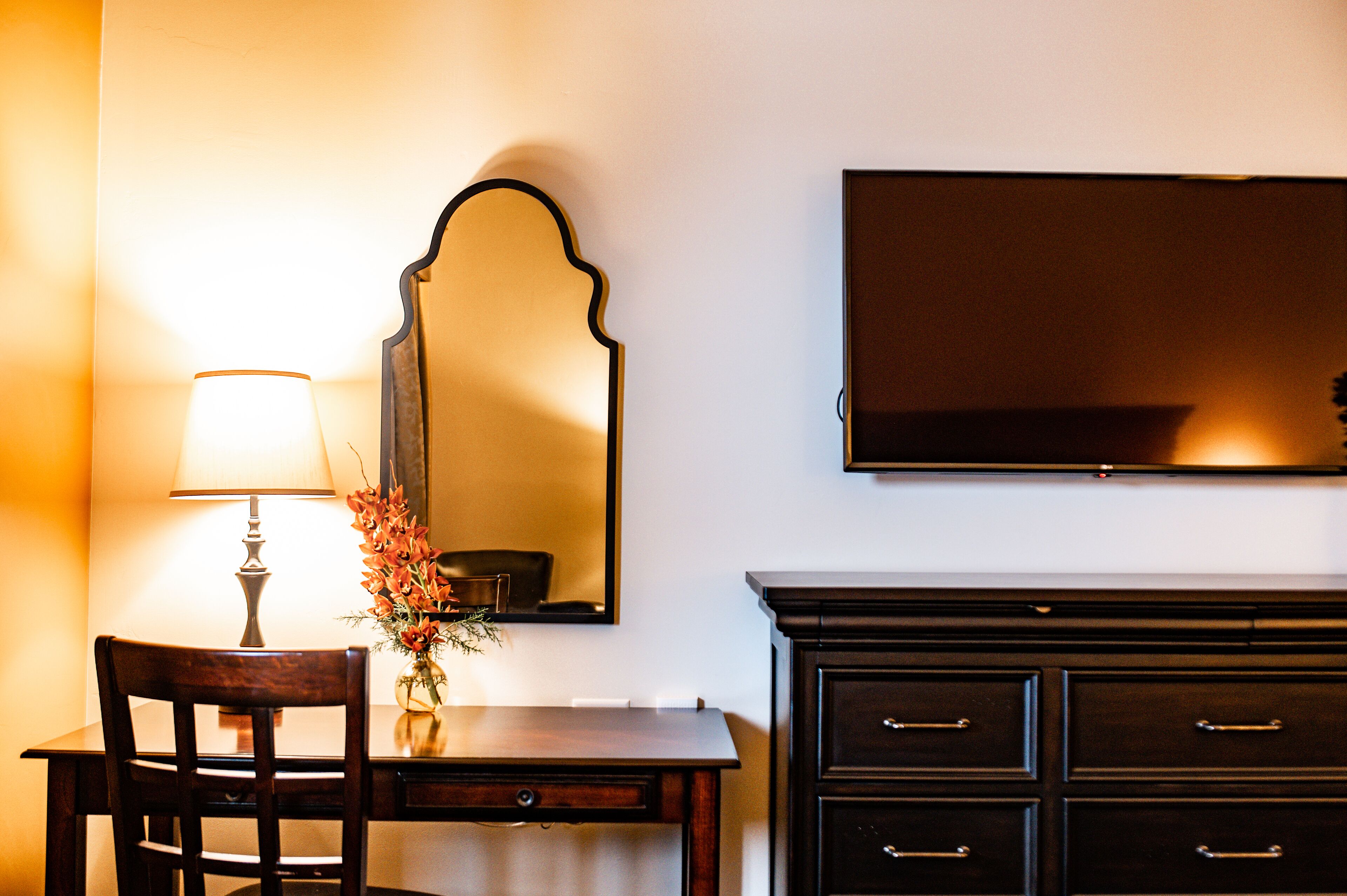 A room with a wooden desk, a lamp, a vase with flowers, a mirror, and a dark-colored dresser.
