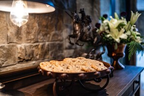 A rustic stone wall with a decorative metal sculpture, a wooden table with a metal tray holding bread, and a vase with flowers.