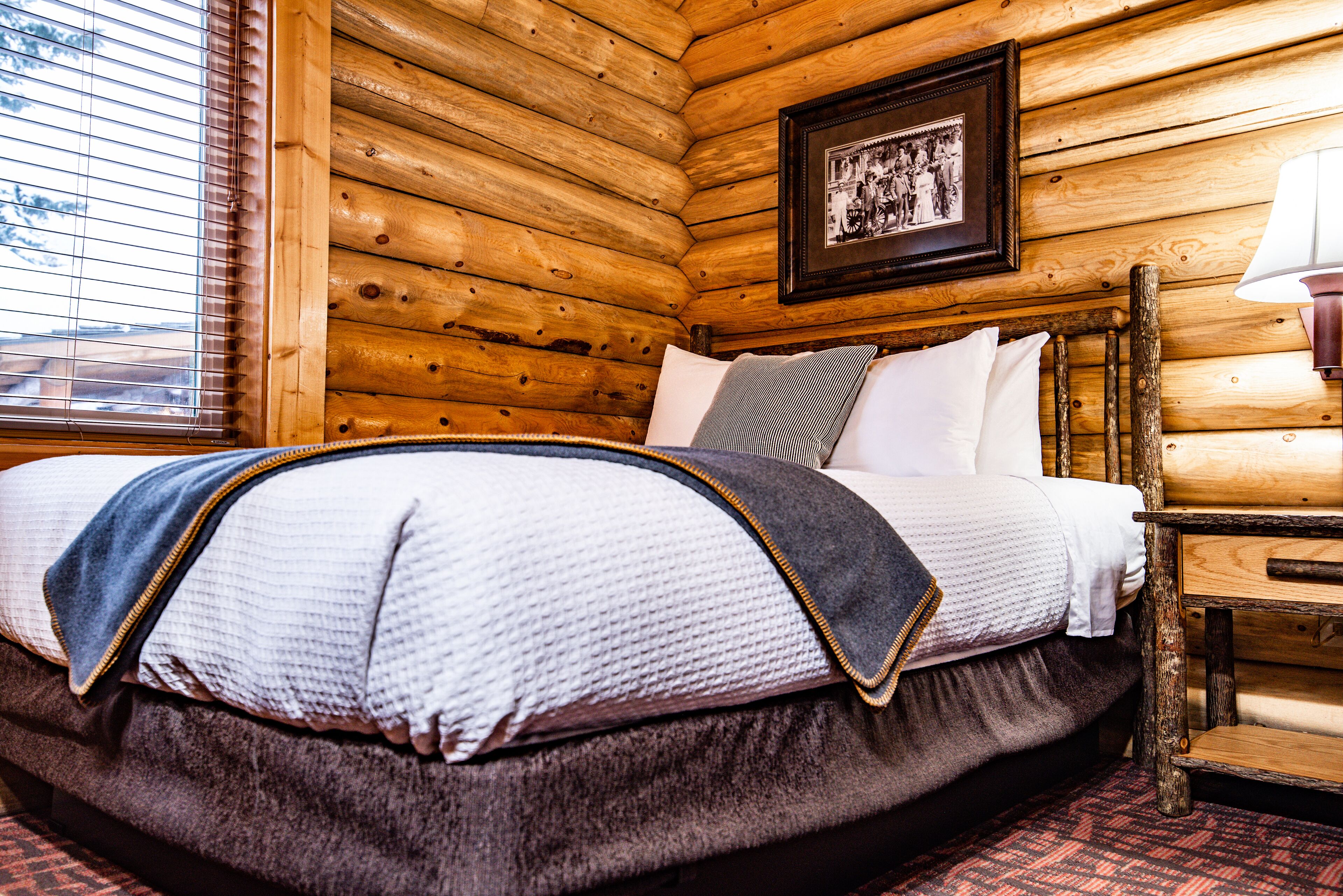A log cabin room with a bed, wooden walls, a framed picture, and a window with blinds.