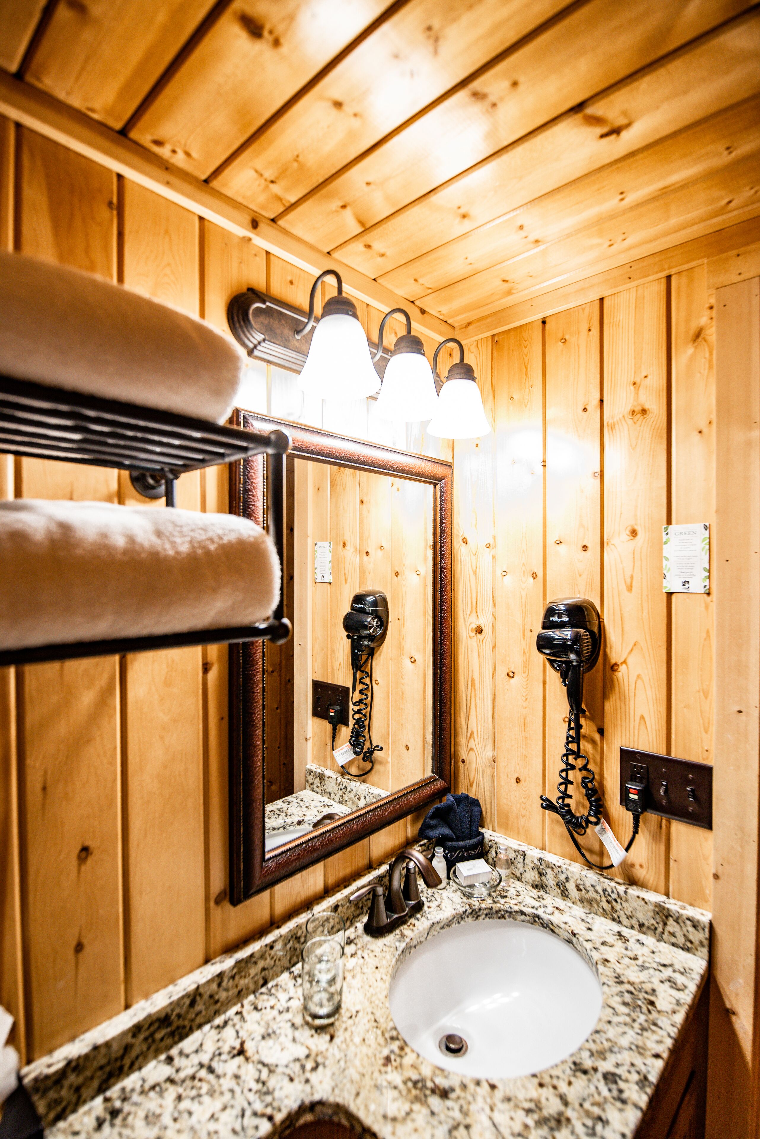 A wooden cabin-style bathroom with a granite countertop sink, a towel rack with towels, a hairdryer, and a mirror.