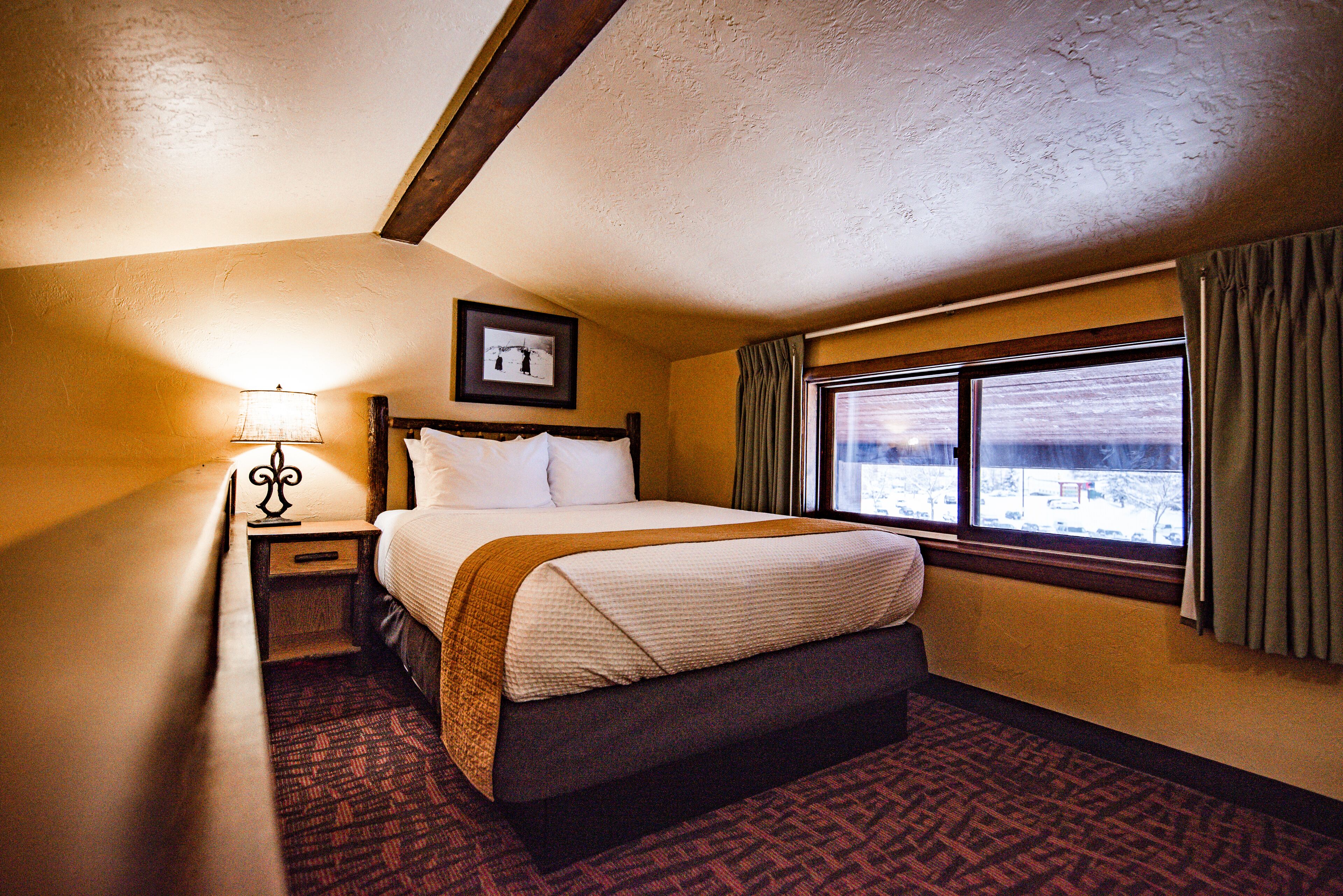 A hotel room with a bed, a nightstand, a lamp, a window with curtains, and a view of the outside.