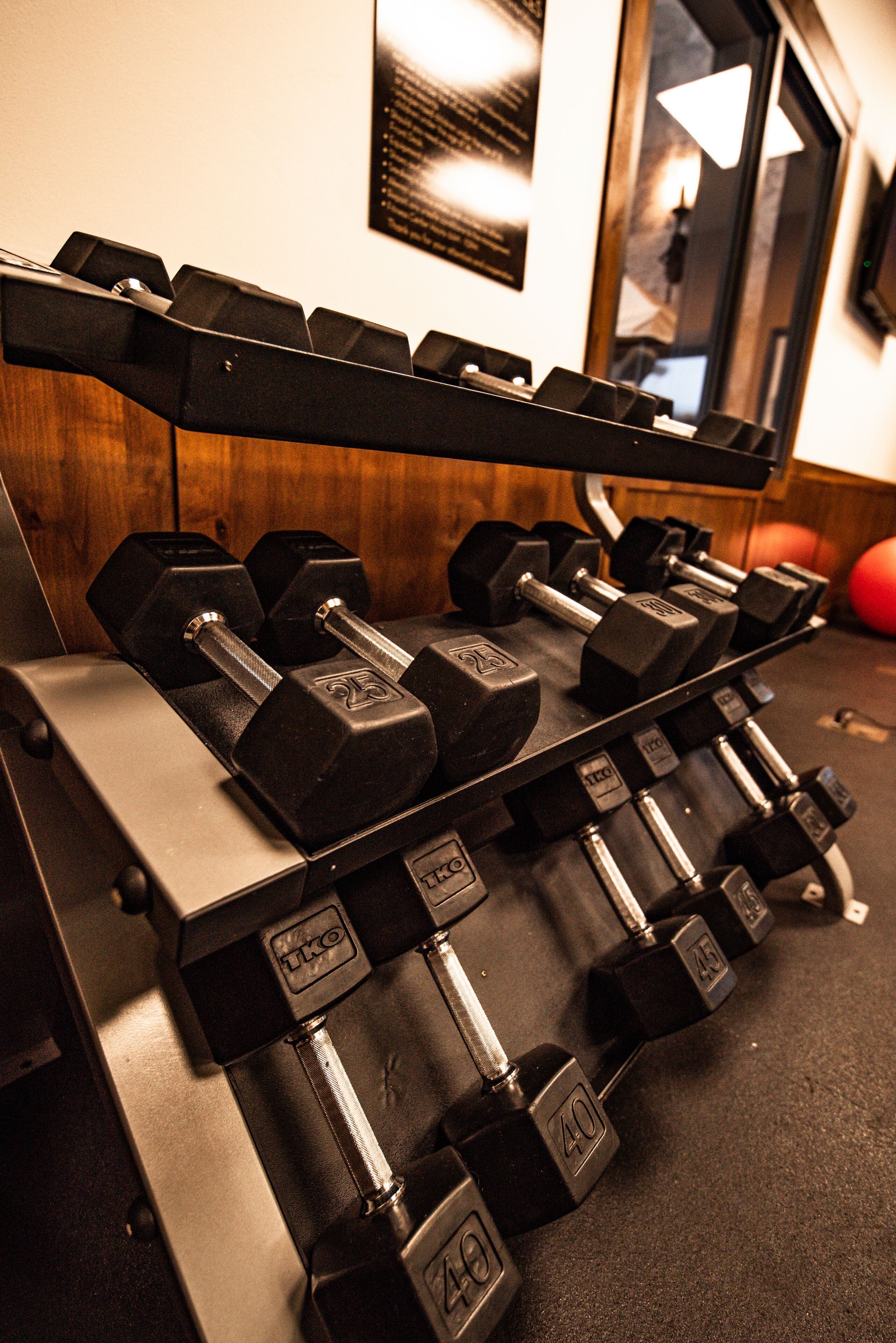 A set of black dumbbells on a rack with weight markings, a wooden wall, and a mirror reflecting a room.