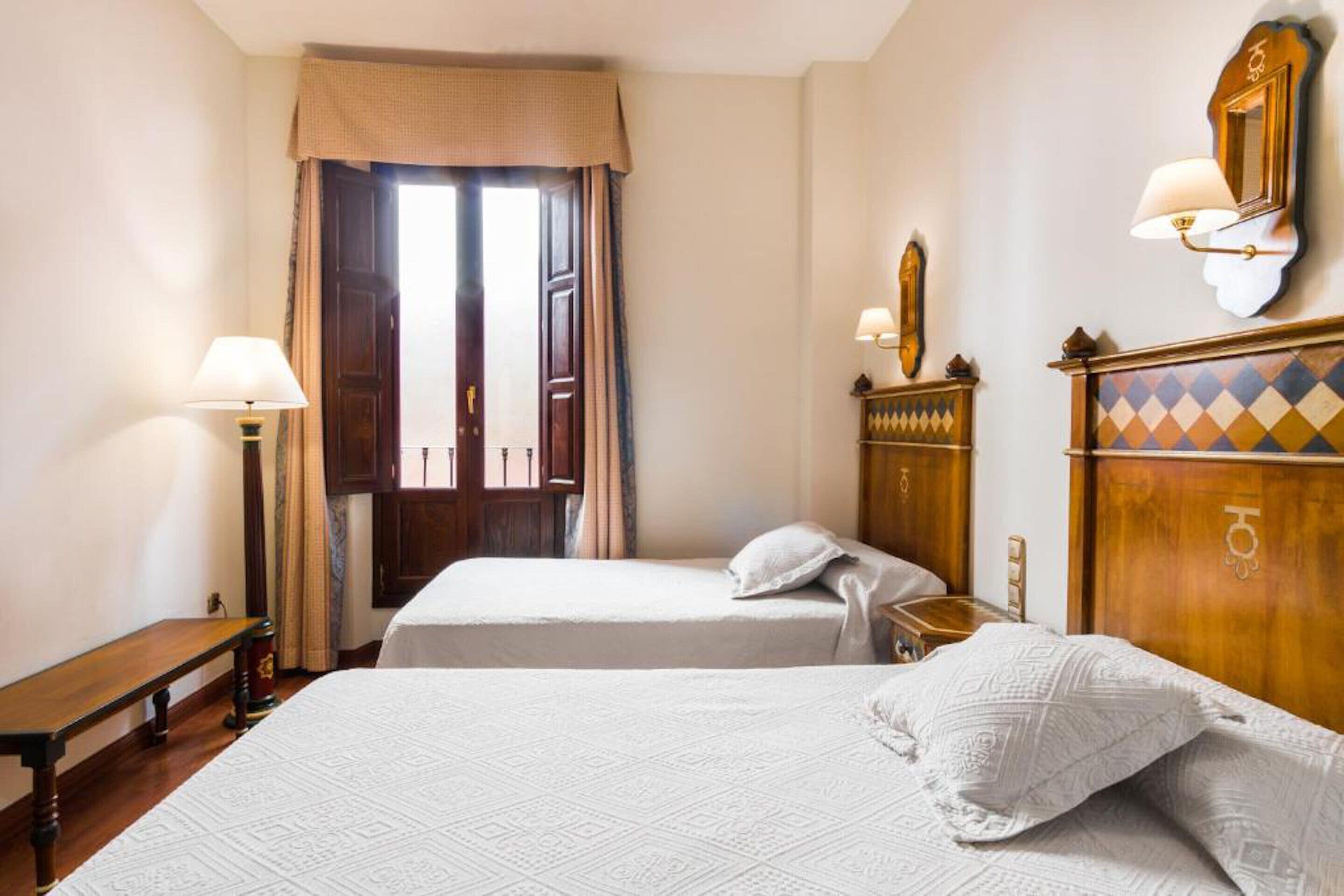 A hotel room with two beds, a bench, a lamp, a mirror, and a window with curtains.