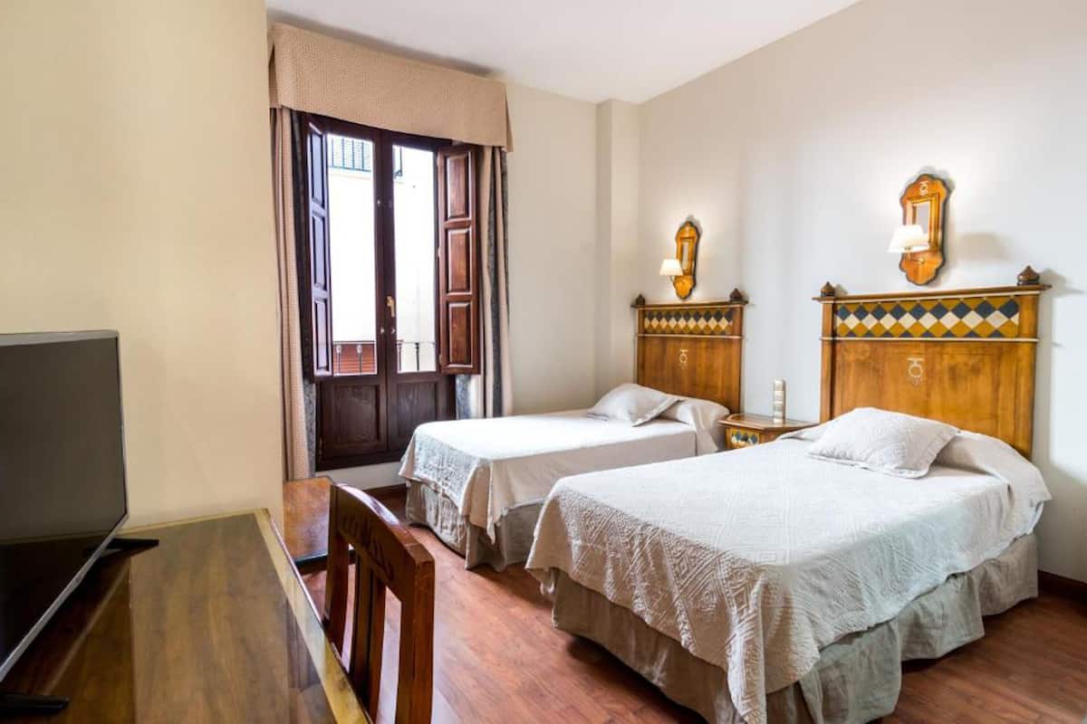 Quarto de hotel com duas camas de solteiro, uma escrivaninha de madeira, televisão e uma janela com cortinas.