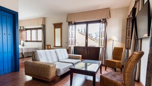 A living room with a sofa, armchairs, a coffee table, and a television. There is a window with curtains and a view of buildings outside.