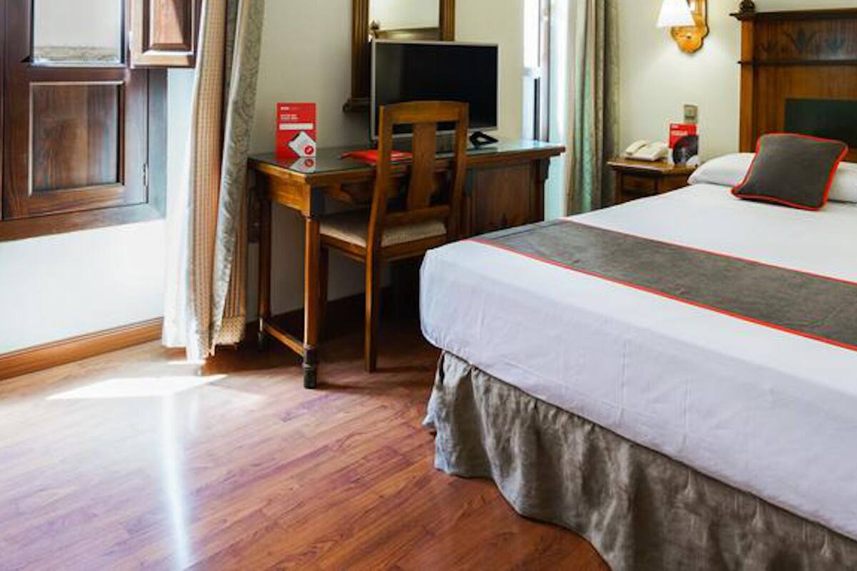Quarto de hotel com cama, escrivaninha, cadeira e janela.