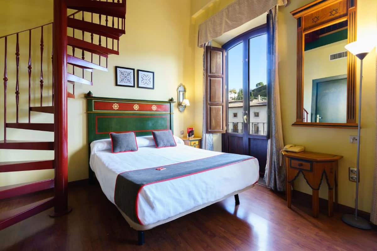 Quarto com escada de madeira, cama com cabeceira verde, mesa de cabeceira, espelho e janela com cortinas.