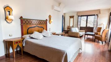 A hotel room with a large bed, a wooden nightstand, a chair, a small table, and a window with a view of buildings.