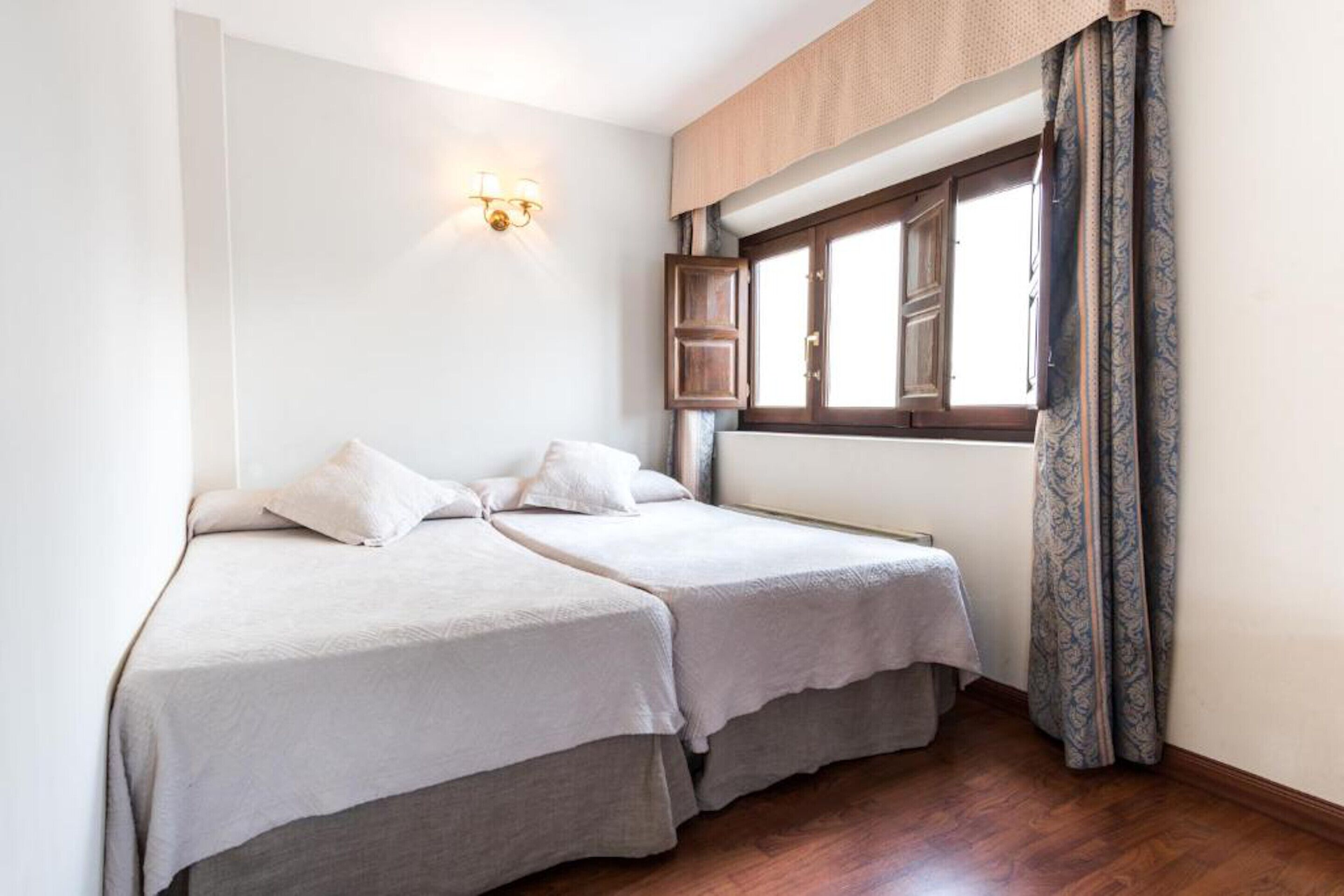 Una habitación con dos camas, una ventana con cortinas y una lámpara fijada a la pared.