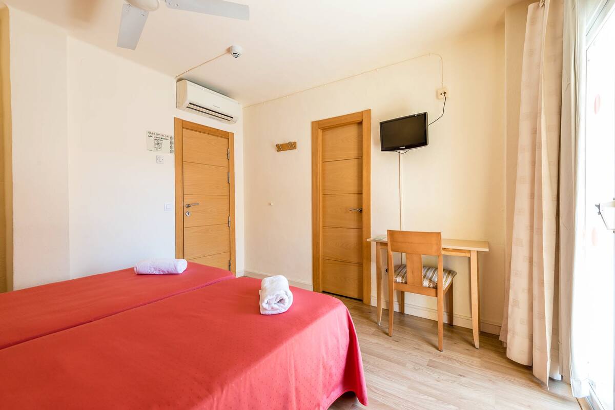 Ein Hotelzimmer mit Bett, Schreibtisch, Stuhl, Klimaanlage, Fernseher und einem Vorhangfenster.