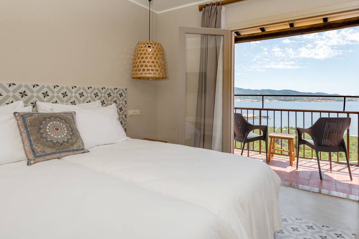 Une chambre avec un grand lit, un coussin à motifs, un luminaire suspendu et un balcon avec des chaises et une table.