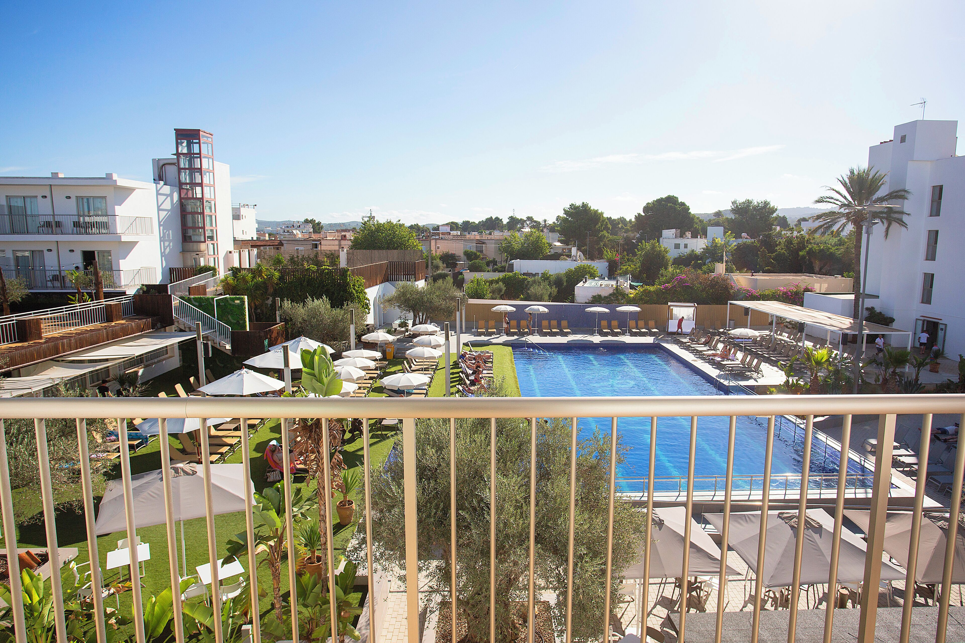 A pool area with sun loungers and umbrellas, surrounded by greenery and buildings.