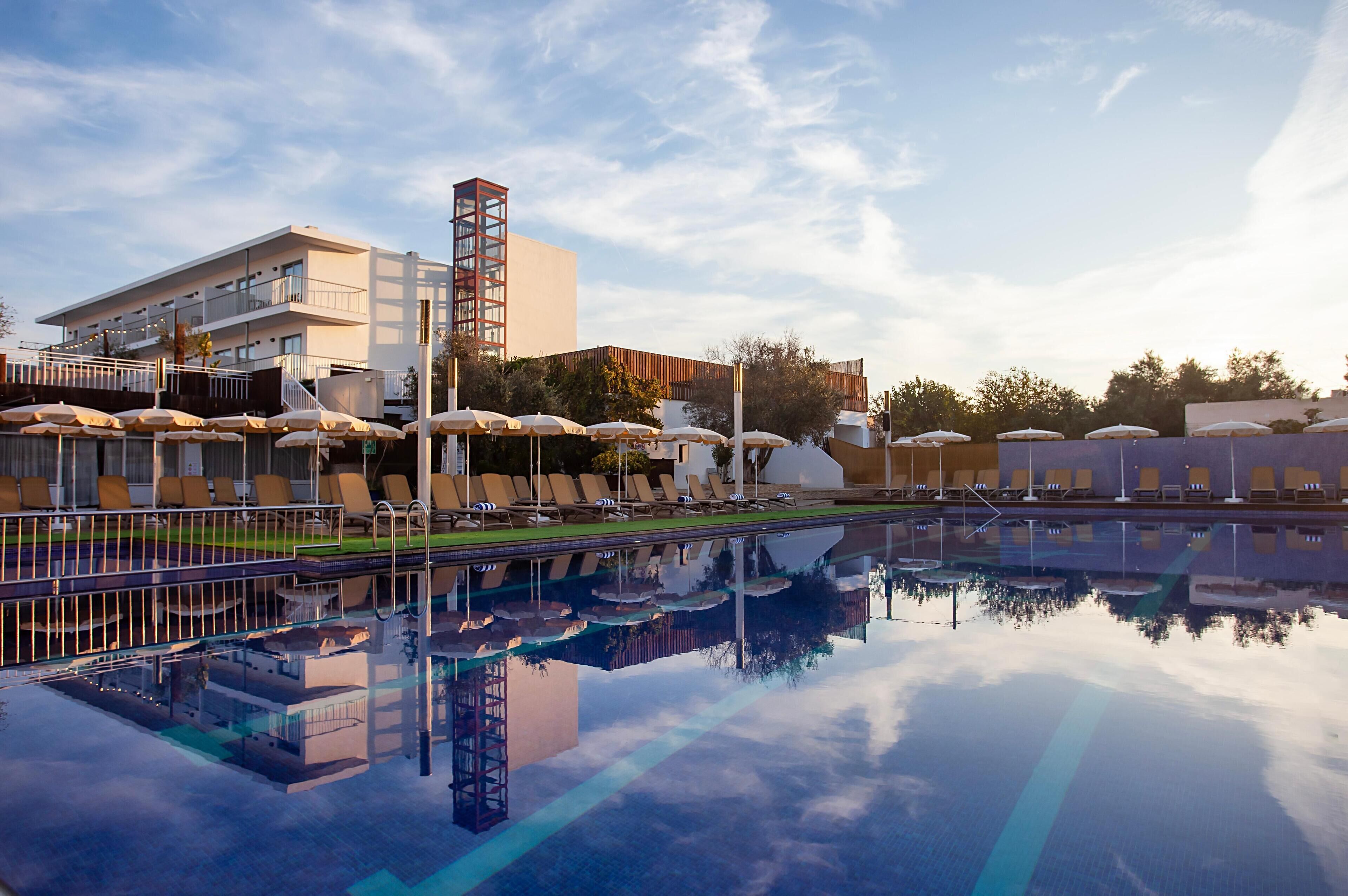 A swimming pool with lounge chairs and umbrellas, a modern building with balconies, and a clear sky.