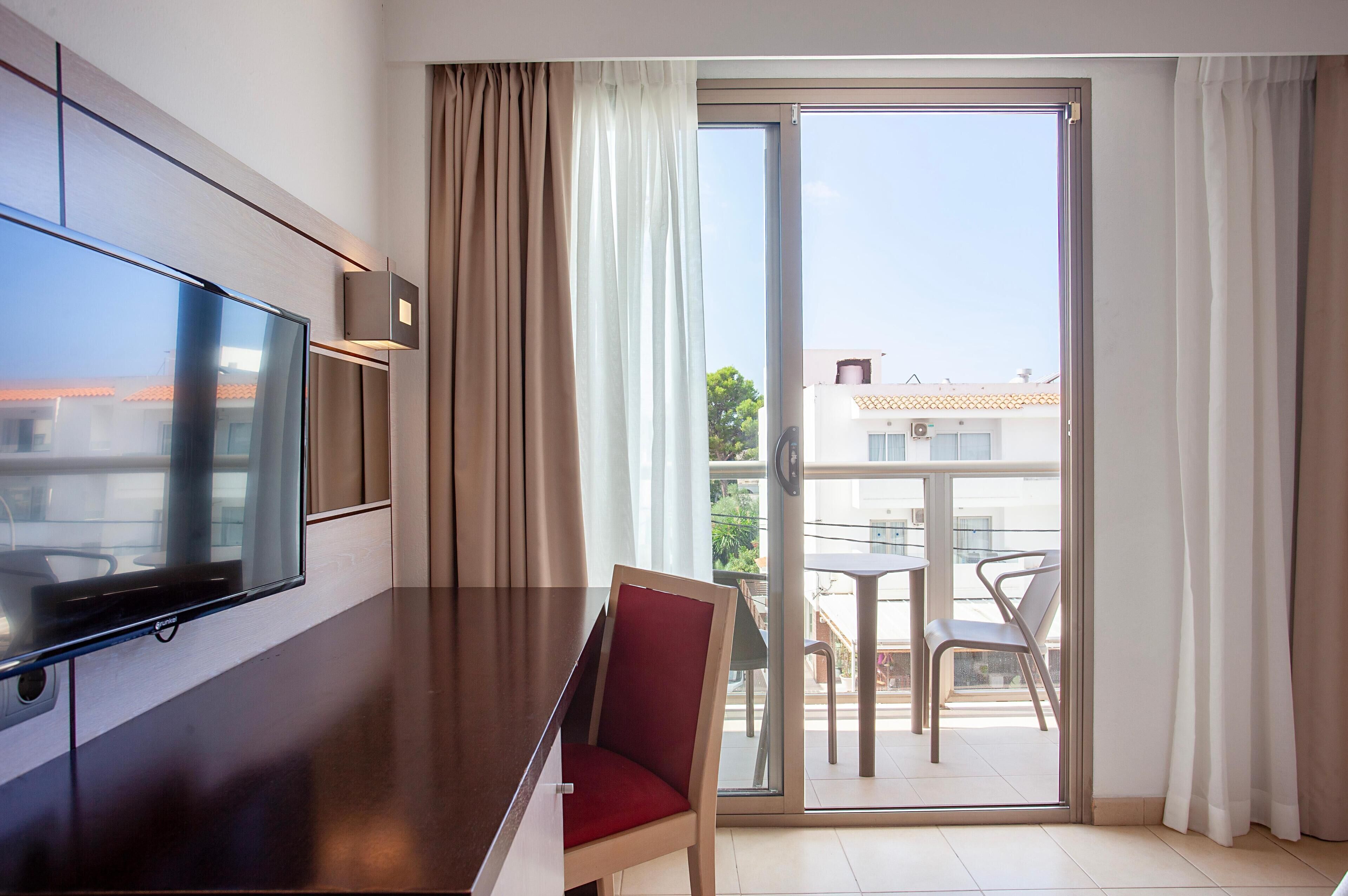 A hotel room with a flat-screen TV mounted on the wall, a wooden dining table, a red chair, and a sliding glass door leading to an outdoor balcony with a table and chairs.