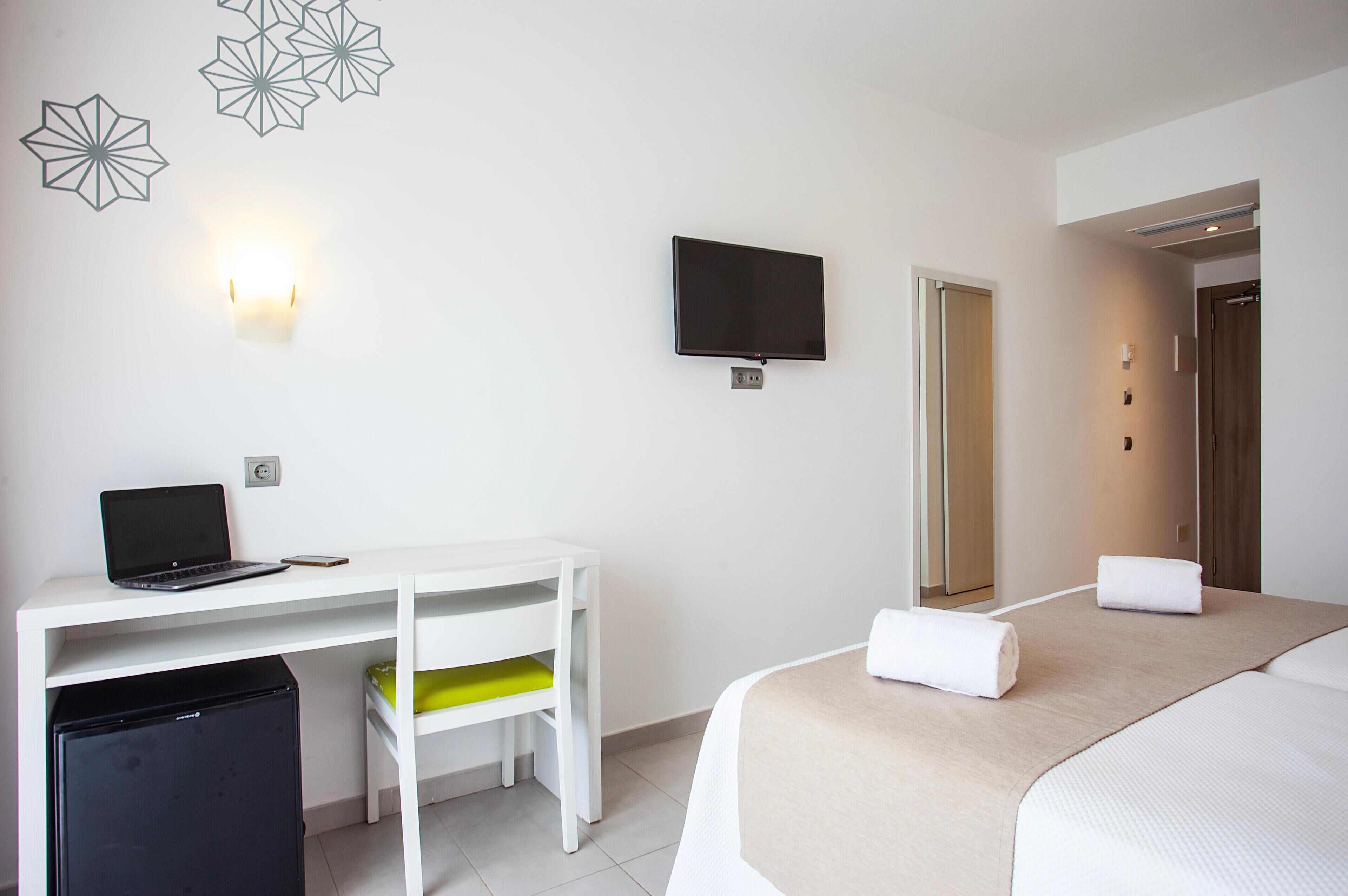 A hotel room with a bed, a desk with a laptop, a TV, and a refrigerator.