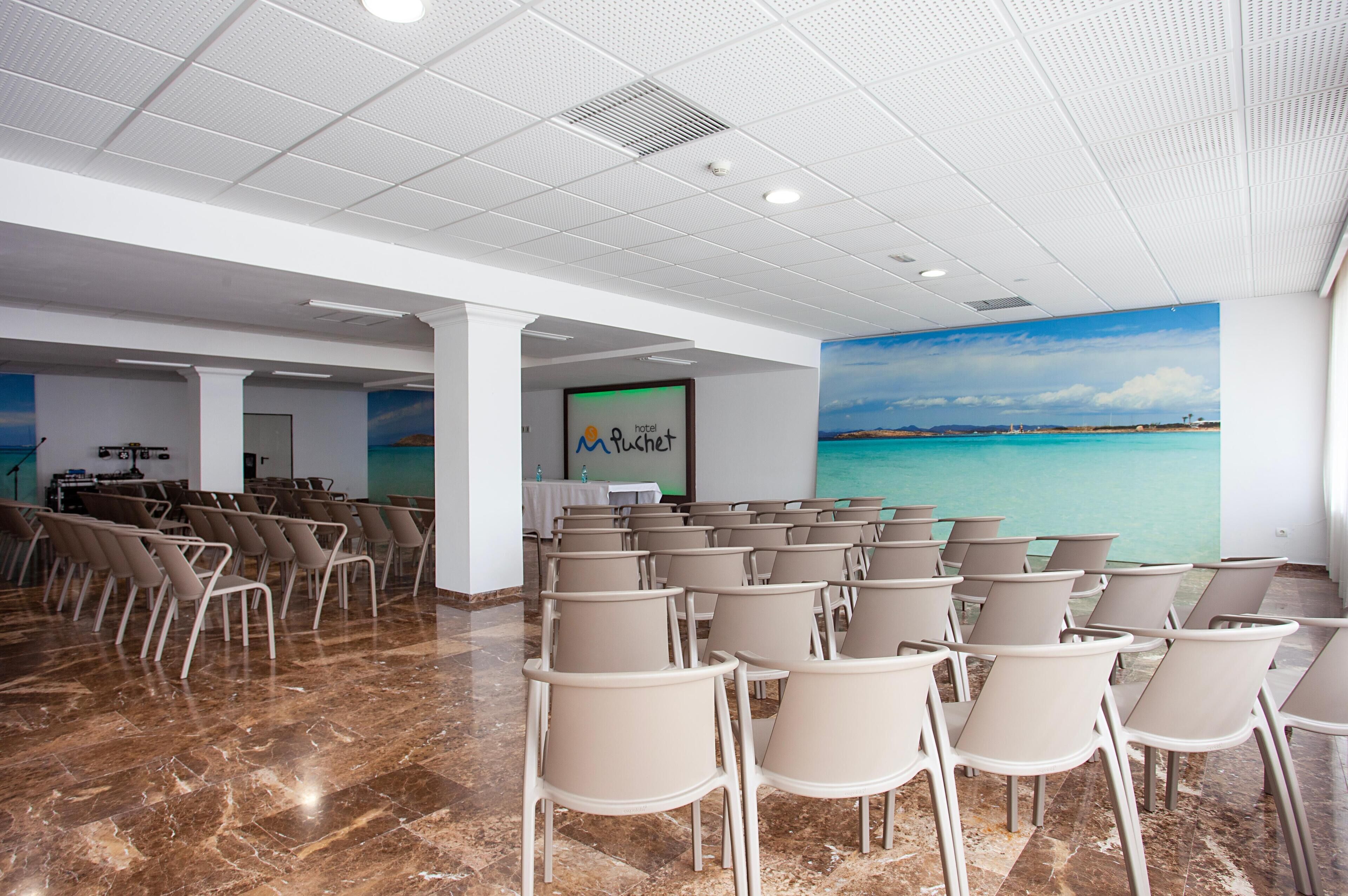 Ein Konferenzraum mit einem großen Bildschirm, beigen Stühlen und einem Wandgemälde mit Strandmotiv.