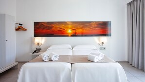Habitación de hotel con un cuadro grande de una puesta de sol, dos camas con sábanas blancas, una mesita de noche con lámpara y un teléfono.