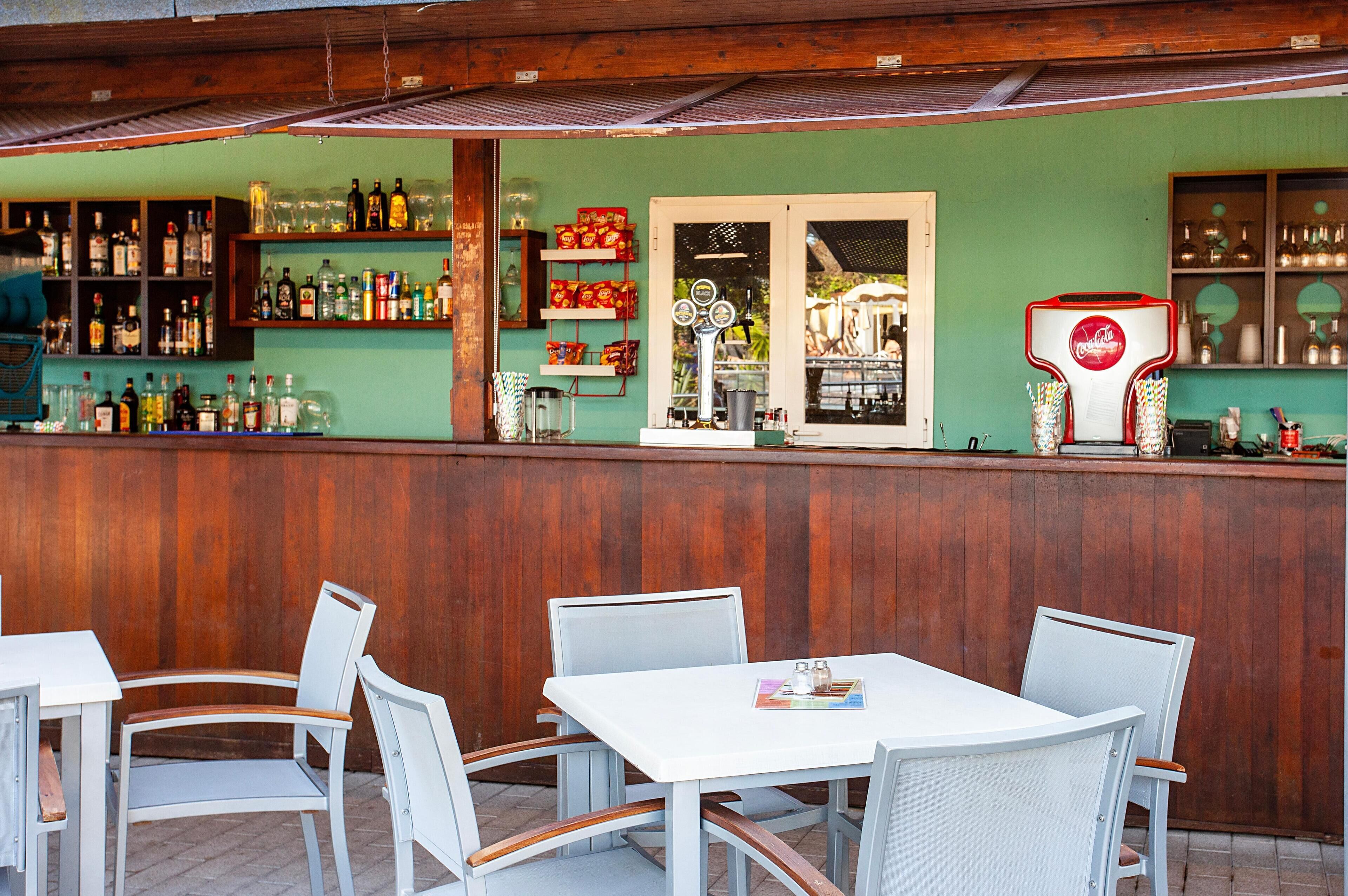 Un espace bar extérieur doté d’un comptoir en bois, de chaises blanches et d’un réfrigérateur arborant la marque Coca-Cola.