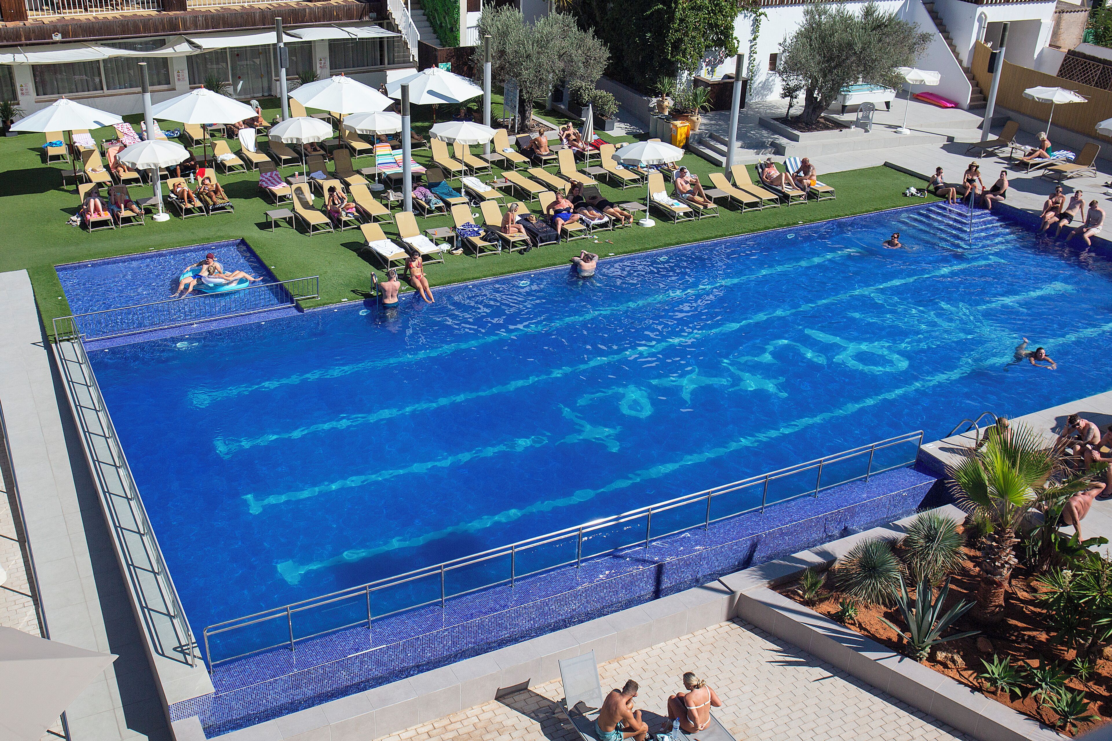 Una amplia piscina con tumbonas y sombrillas, rodeada de un área ajardinada con plantas.