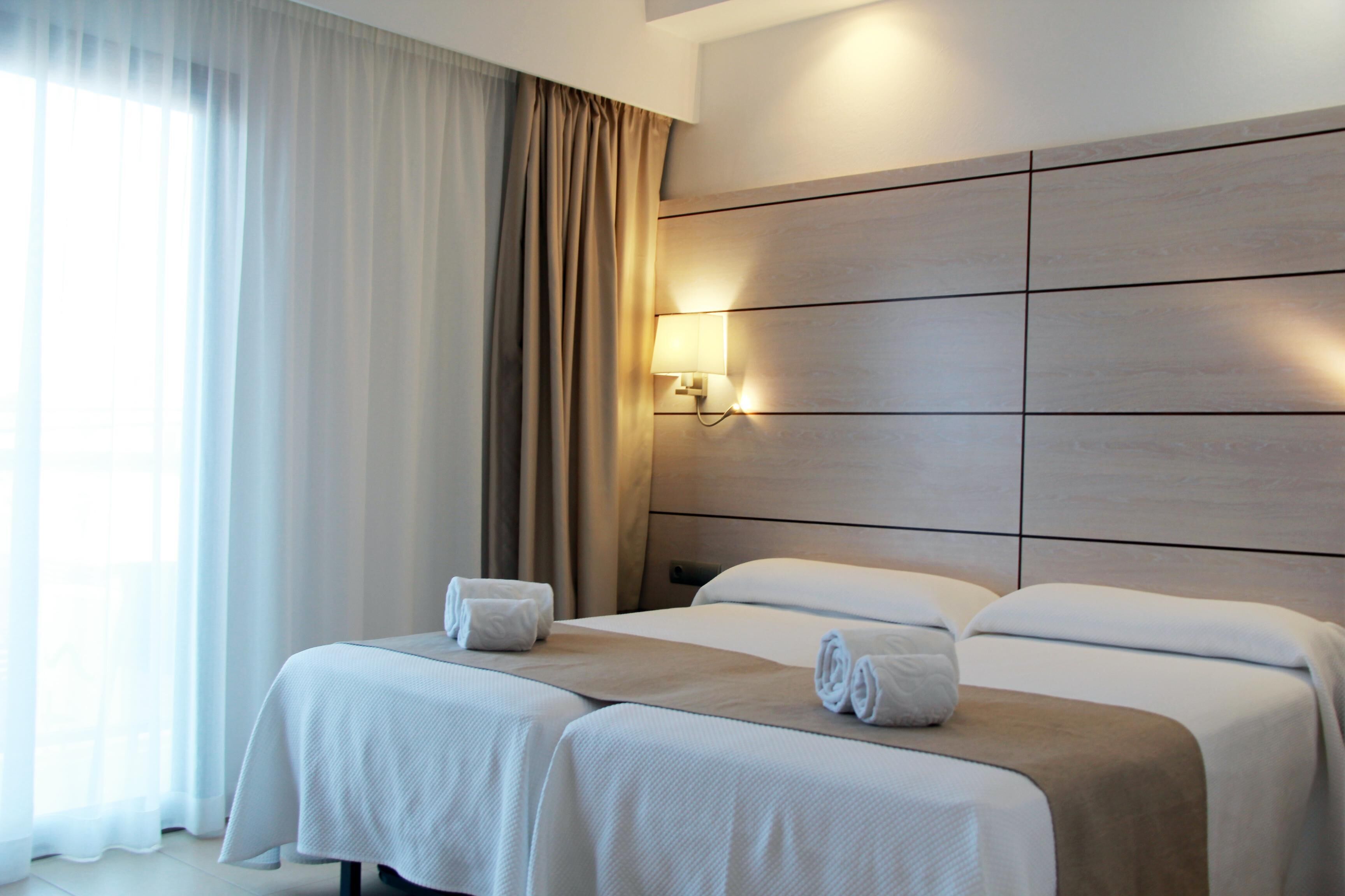 Een hotelkamer met twee bedden, een groot raam met gordijnen, een wandlamp en een houten hoofdeinde.