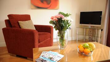 A living room with a red armchair, a glass coffee table with magazines and fruit, a vase with flowers, and a TV on a stand.